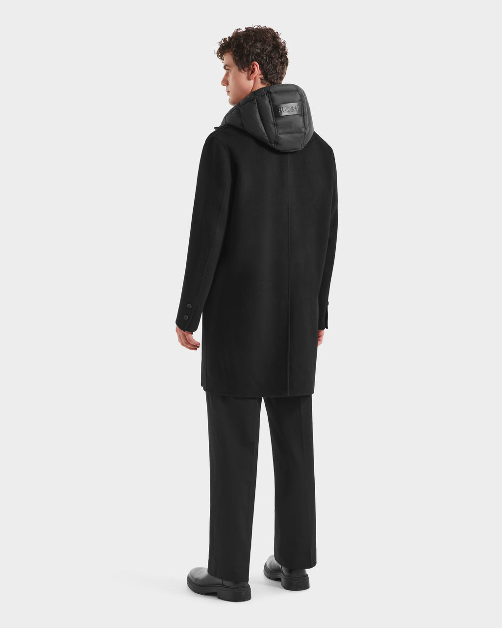 A person with short curly hair stands facing away, wearing the Rudsak JEREMIAH - 6125509 Black men’s wool-blend coat with a hood and black patch, paired with black pants and shoes against a plain white background.