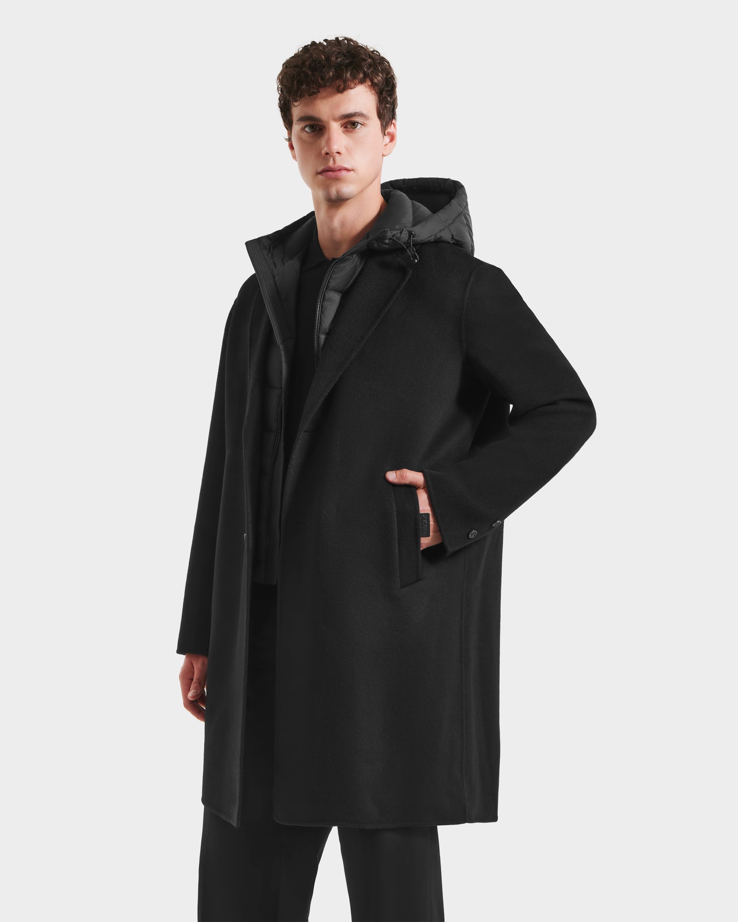 A man with short curly hair wears the Rudsak JEREMIAH - 6125509 Black wool-blend hooded coat over a black outfit, standing with one hand in his pocket against a white background—a modern cold-weather essential.