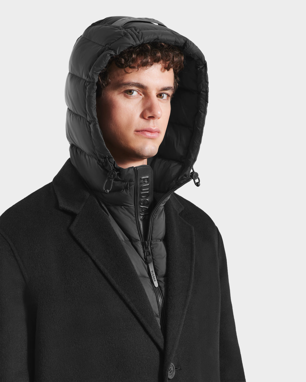 A young man with curly hair models the Rudsak JEREMIAH - 6125509 Black men’s wool-blend coat, which features a down-insulated inner dickie, posing against a plain light background—a modern cold-weather essential.
