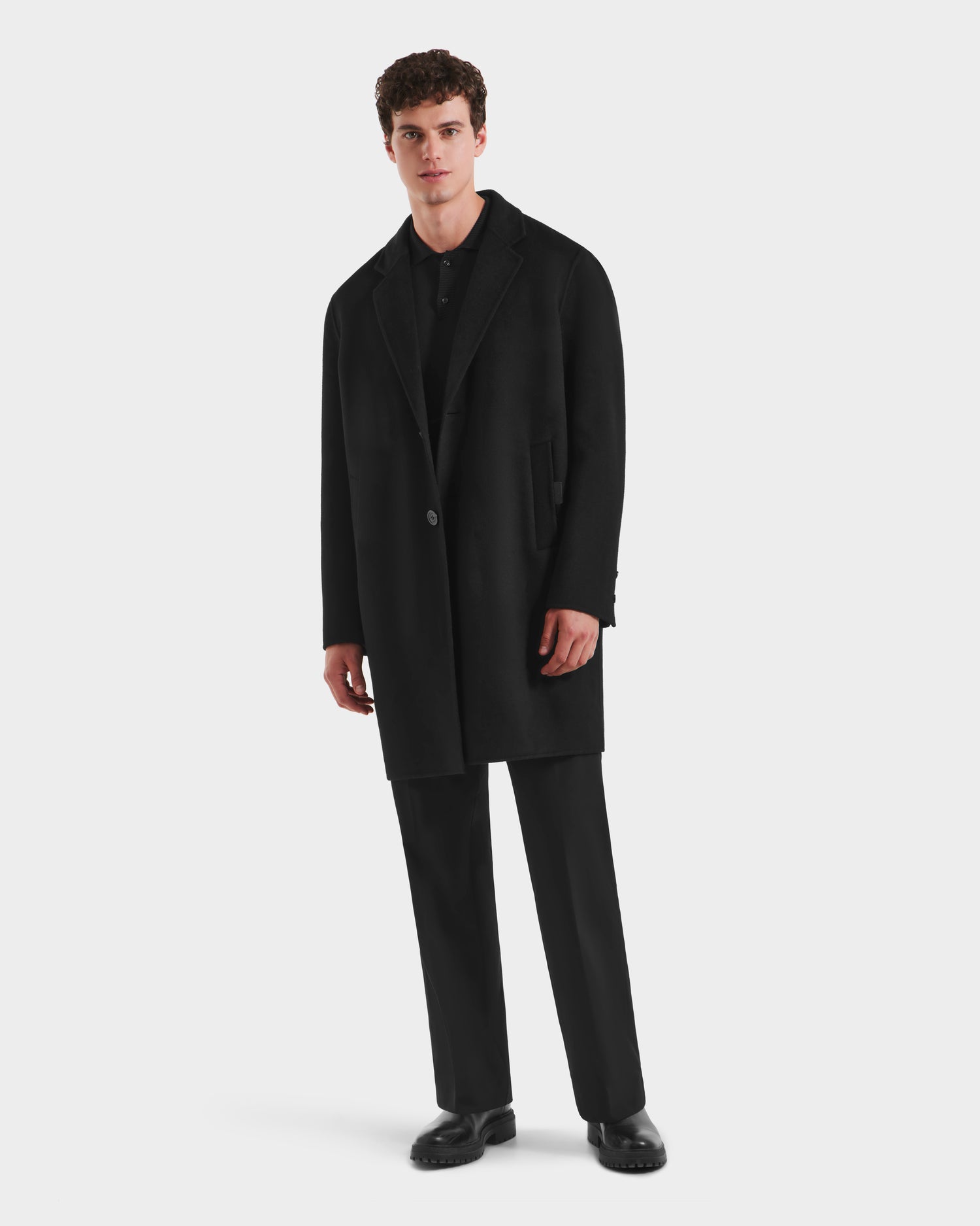 A man stands against a white background wearing the Rudsak JEREMIAH - 6125509 Black men’s wool-blend coat with a down-insulated inner dickie, paired with black shirt, trousers, and shoes—a true cold-weather staple.
