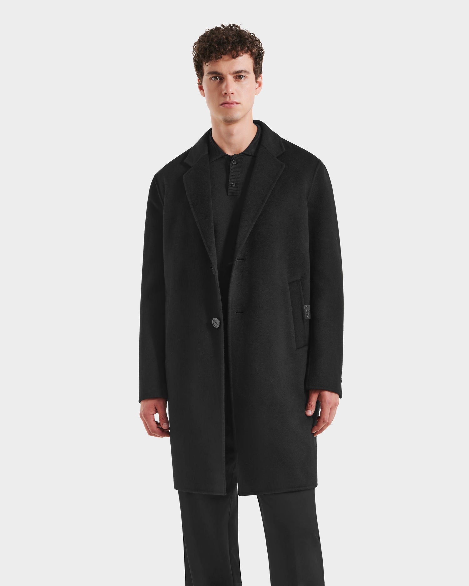 A young man with curly hair models the Rudsak JEREMIAH - 6125509 Black wool-blend coat over a black shirt and black pants, standing against a plain white background.