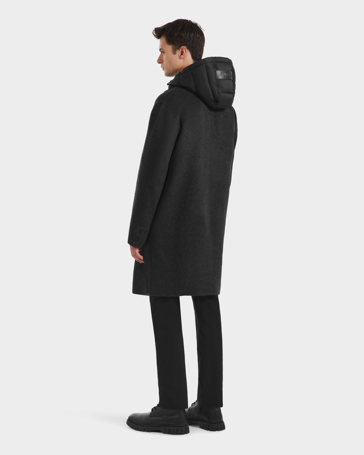 A man faces away, wearing the Rudsak JEREMIAH - 6125509 Grey wool-blend hooded coat with a removable down-insulated dickie, paired with black pants and shoes, against a plain white background.