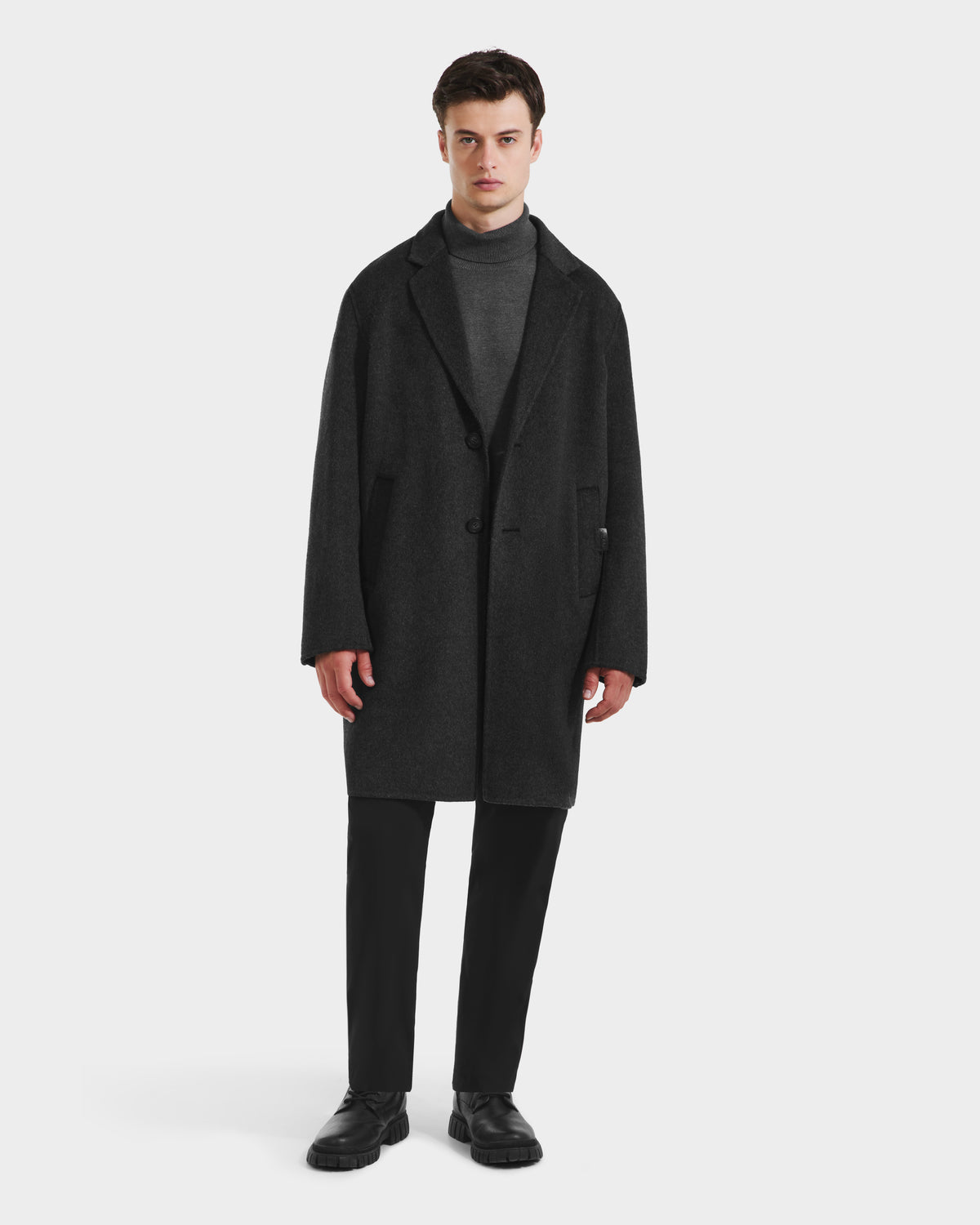 A man stands facing forward wearing the Rudsak JEREMIAH - 6125509 Grey men’s wool-blend coat over a gray turtleneck, black pants, and black shoes—a refined cold-weather staple—against a plain white background.