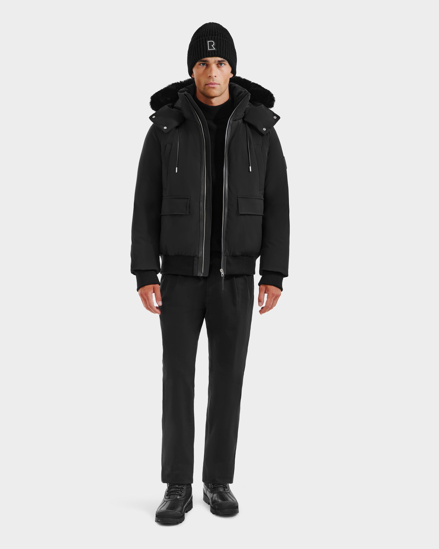 A man faces forward wearing the Rudsak JEROME - 6125514 Black men’s down bomber jacket, black beanie, black pants, and black shoes against a plain white background.