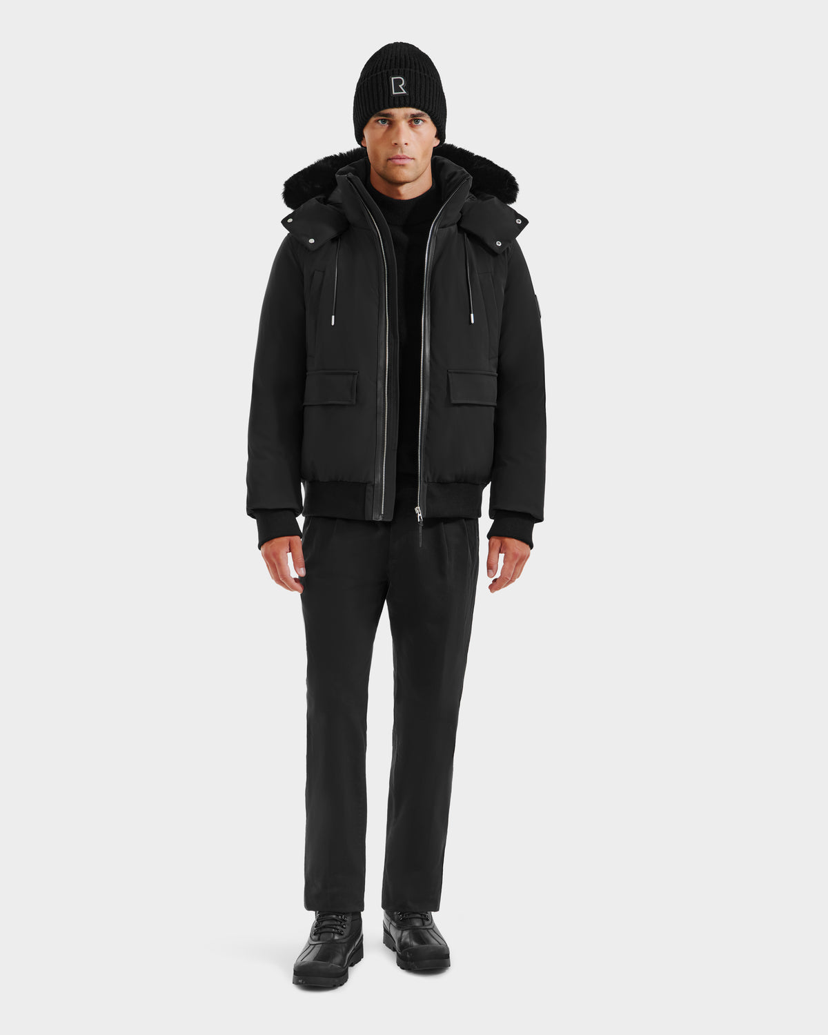 A man faces forward wearing the Rudsak JEROME - 6125514 Black men’s down bomber jacket, black beanie, black pants, and black shoes against a plain white background.