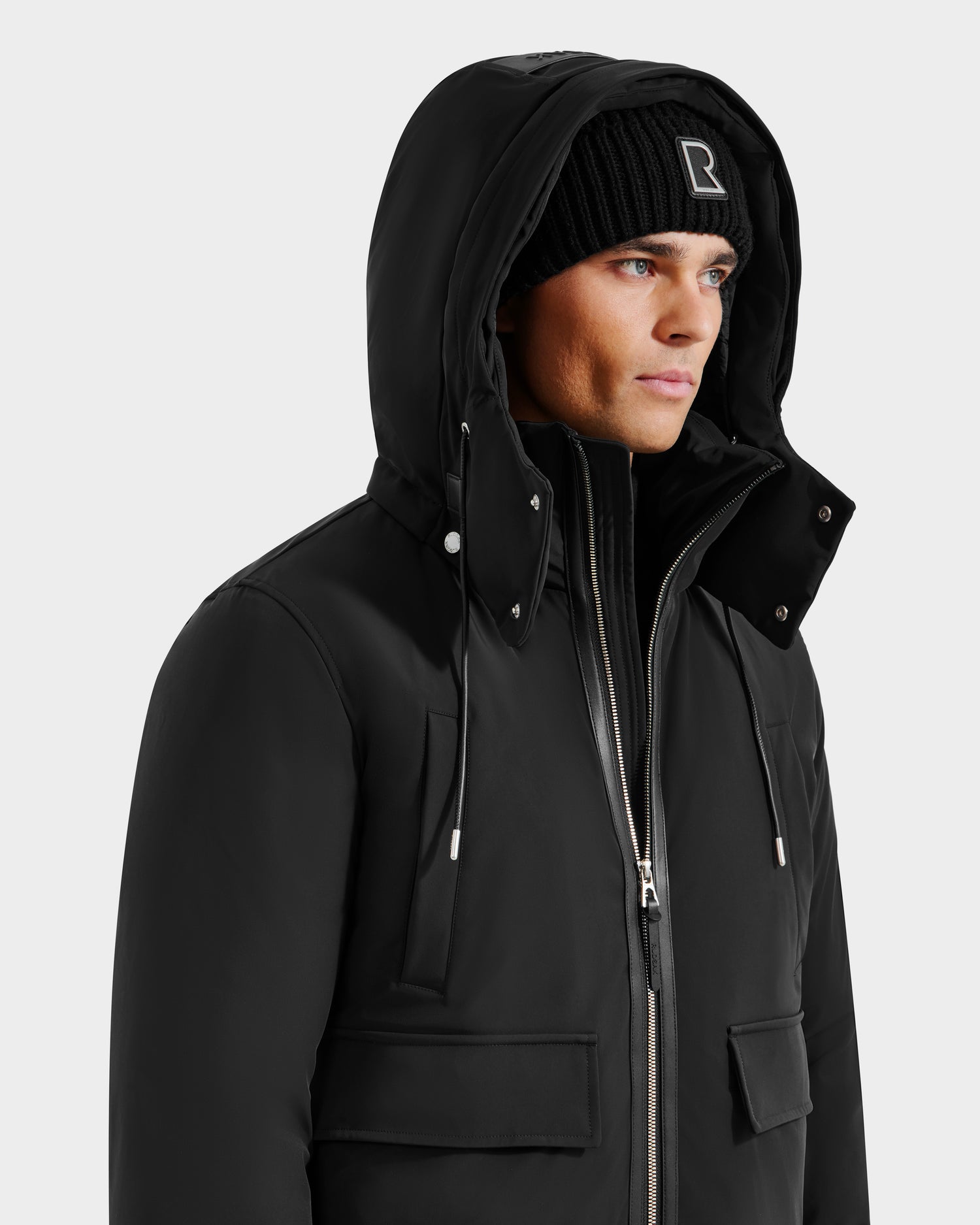 A person wears the Rudsak JEROME - 6125514 Black men’s down bomber jacket with a zipper and flap pockets, paired with a black knit beanie featuring a white letter R, looking to the side against a plain background.