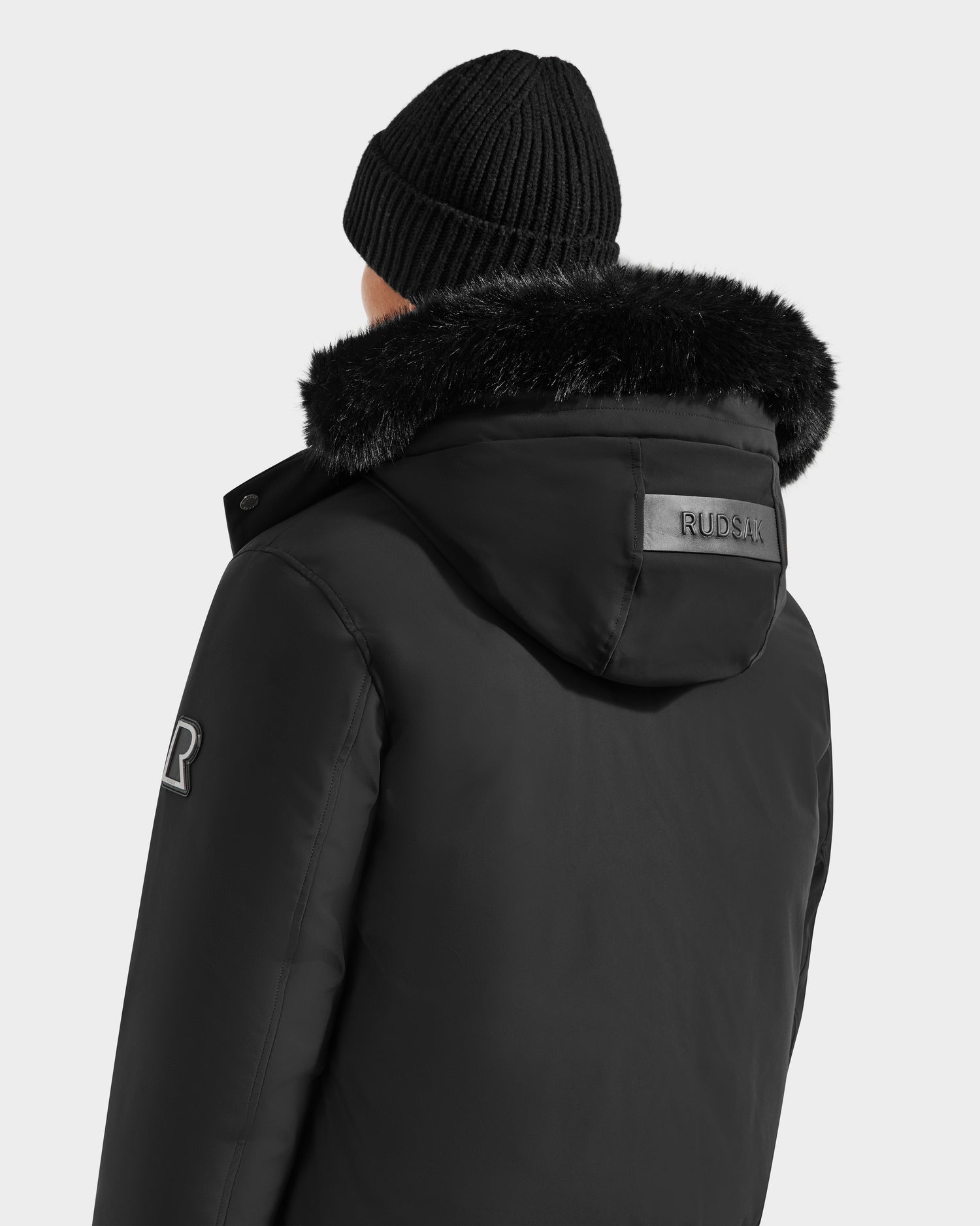 A person wearing the Rudsak JEROME - 6125514 Black beanie and winter performance jacket with faux fur-lined hood, featuring a RUDSAK patch on the hood and R logo on the shoulder, crafted with RDS-certified down. Seen from behind.
