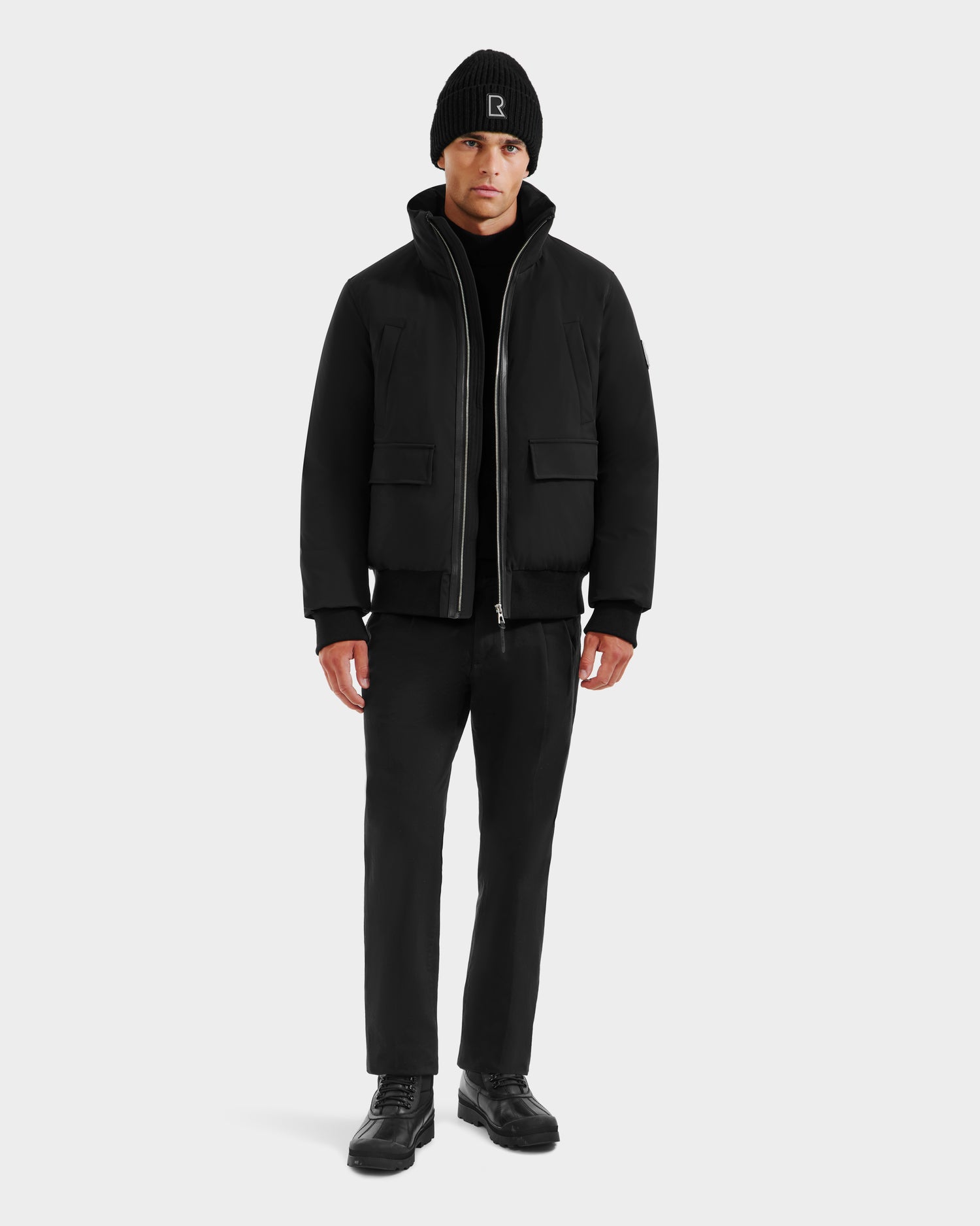 A man faces forward wearing the Rudsak JEROME - 6125514 Black winter performance jacket with front pockets, along with a black beanie, pants, and shoes, against a plain light gray background.