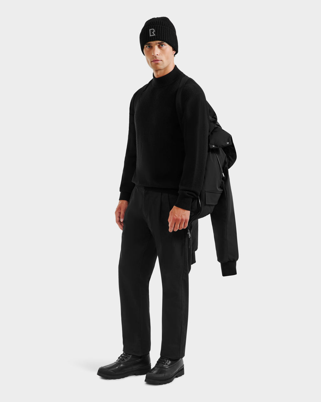 A man in black attire stands holding the Rudsak JEROME - 6125514 Black men’s down bomber jacket and a backpack over his shoulder against a plain light background.
