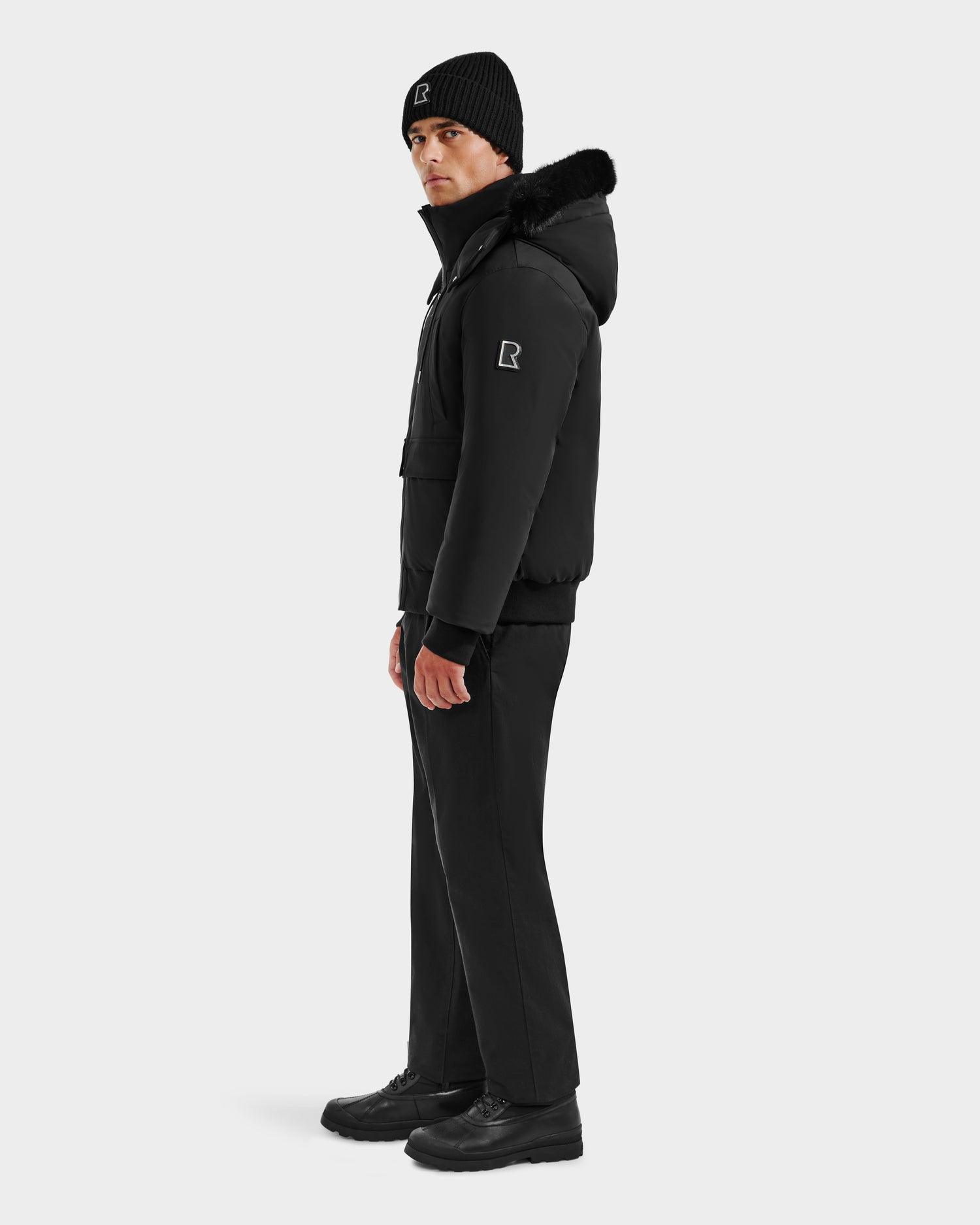 A man stands facing slightly right, wearing the Rudsak JEROME - 6125514 Black men’s down bomber with fur-lined hood, black pants, boots, and a matching R logo on both the jacket sleeve and black beanie.