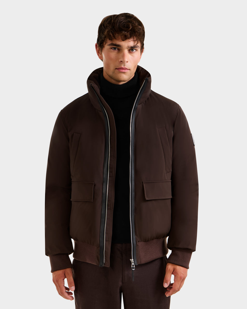 A man wears the JEROME - 6125514 Espresso down bomber jacket by Rudsak, featuring a high collar and front zipper, layered over a black turtleneck. He stands facing forward against a plain, light background.