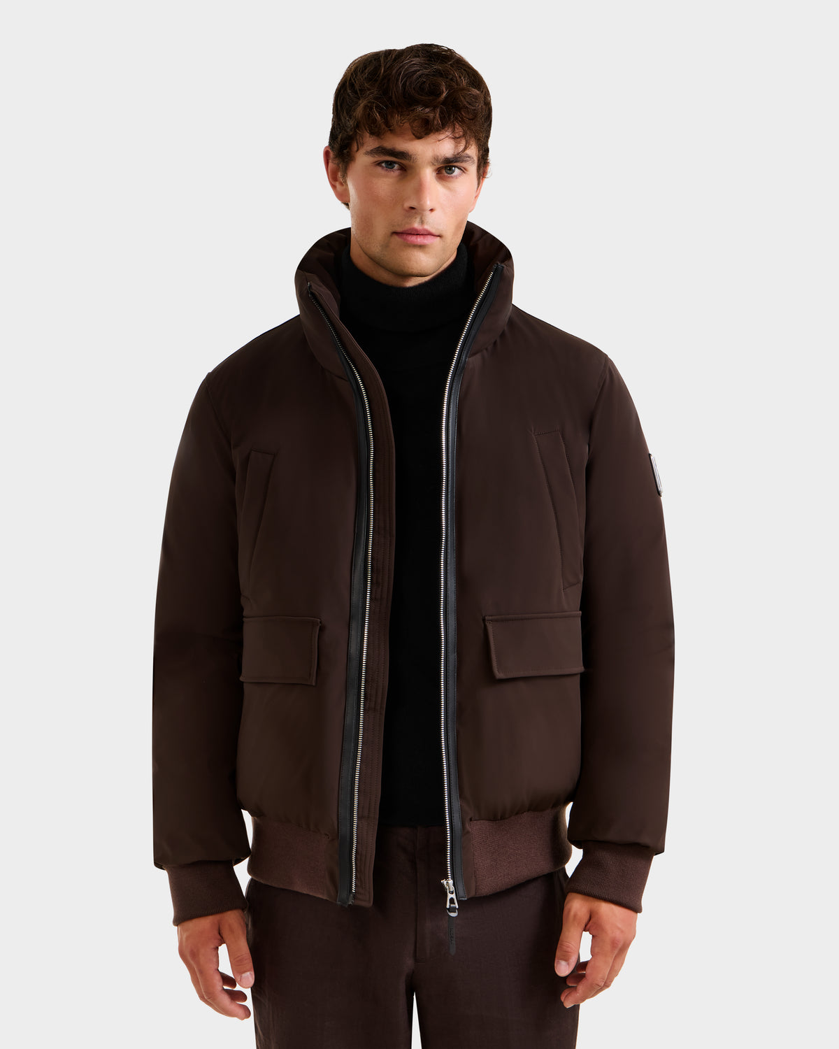 A man wears the JEROME - 6125514 Espresso down bomber jacket by Rudsak, featuring a high collar and front zipper, layered over a black turtleneck. He stands facing forward against a plain, light background.