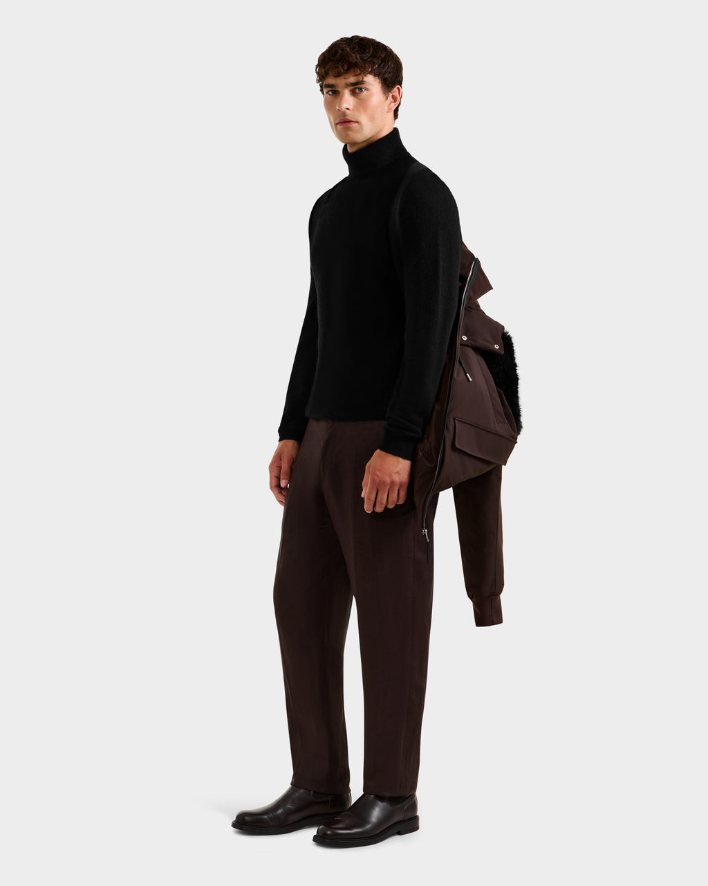 A man stands against a plain background in a black turtleneck, dark brown pants, and black shoes, holding the Rudsak JEROME - 6125514 Espresso RDS-certified down winter performance jacket over one shoulder.