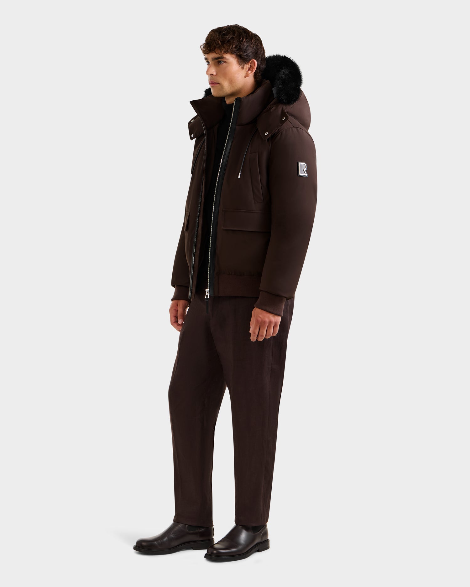 A man wears the Rudsak JEROME - 6125514 Espresso men’s down bomber jacket with a fur-lined hood, brown pants, and black shoes against a plain background—features RDS-certified down for winter performance.