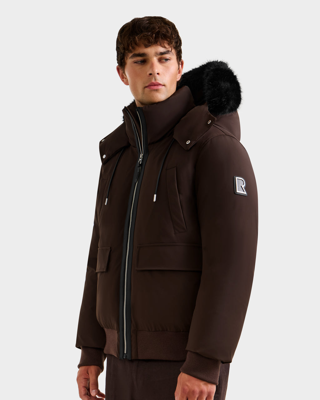 A young man stands against a plain background wearing the Rudsak JEROME - 6125514 Espresso, a men’s down bomber jacket with a black fur-trimmed hood, zippered pockets, RDS-certified down, and an R patch on the upper sleeve.