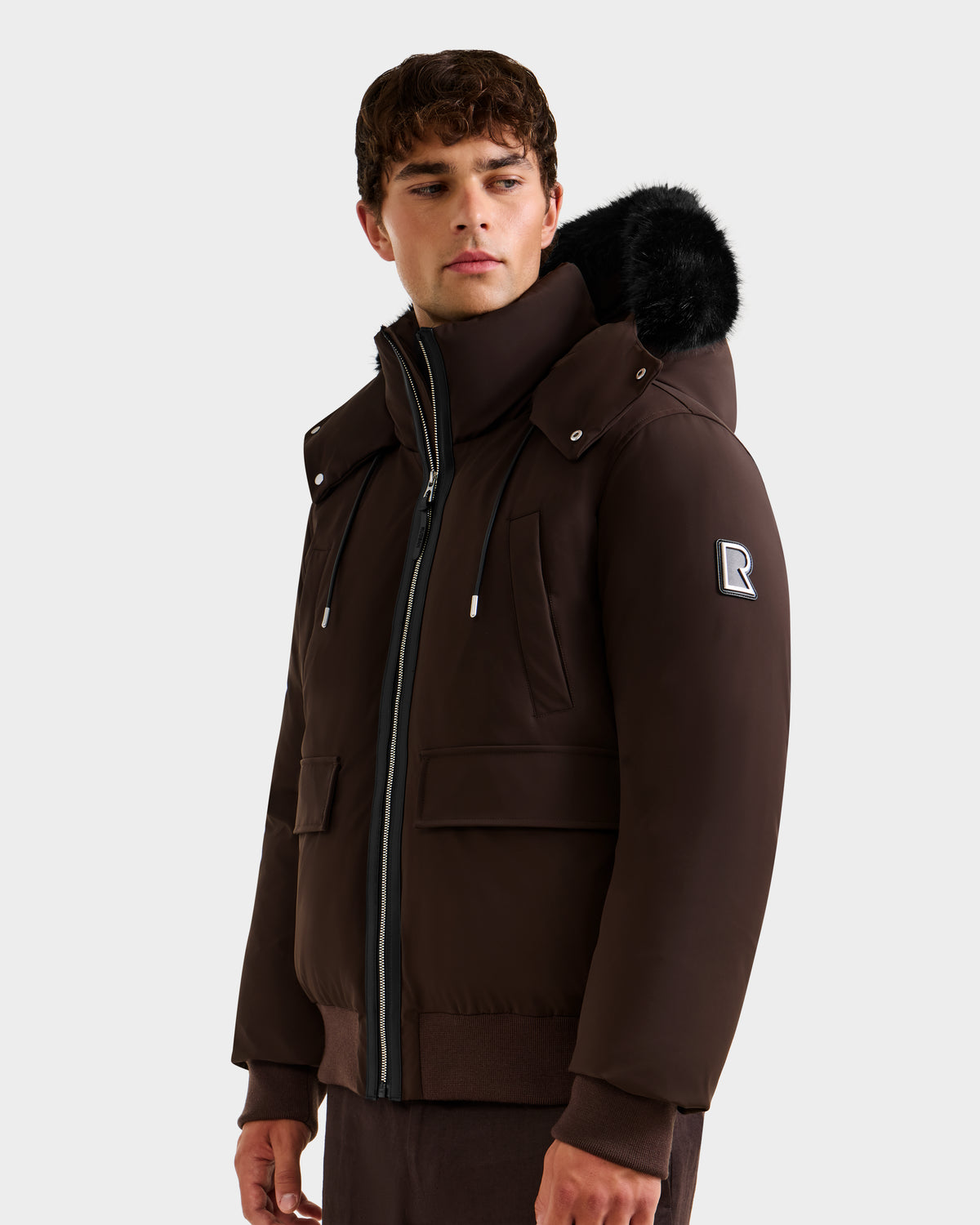 A young man stands against a plain background wearing the Rudsak JEROME - 6125514 Espresso, a men’s down bomber jacket with a black fur-trimmed hood, zippered pockets, RDS-certified down, and an R patch on the upper sleeve.