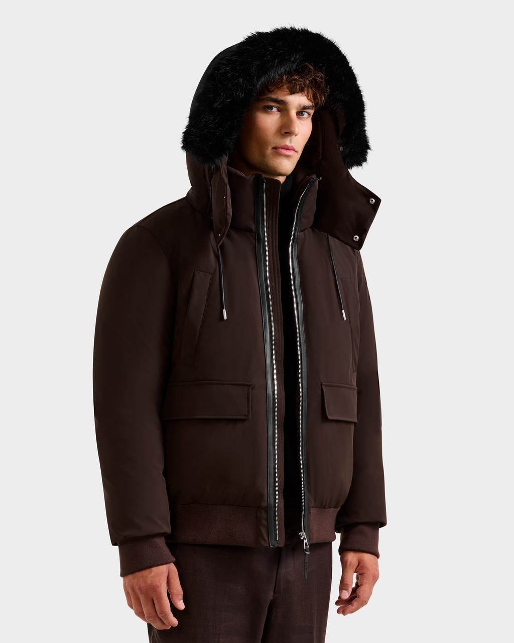 A man wears the Rudsak JEROME - 6125514 Espresso down bomber jacket with black fur trim, front zipper, and flap pockets over brown pants—an ideal winter choice for warmth and style against a light background.