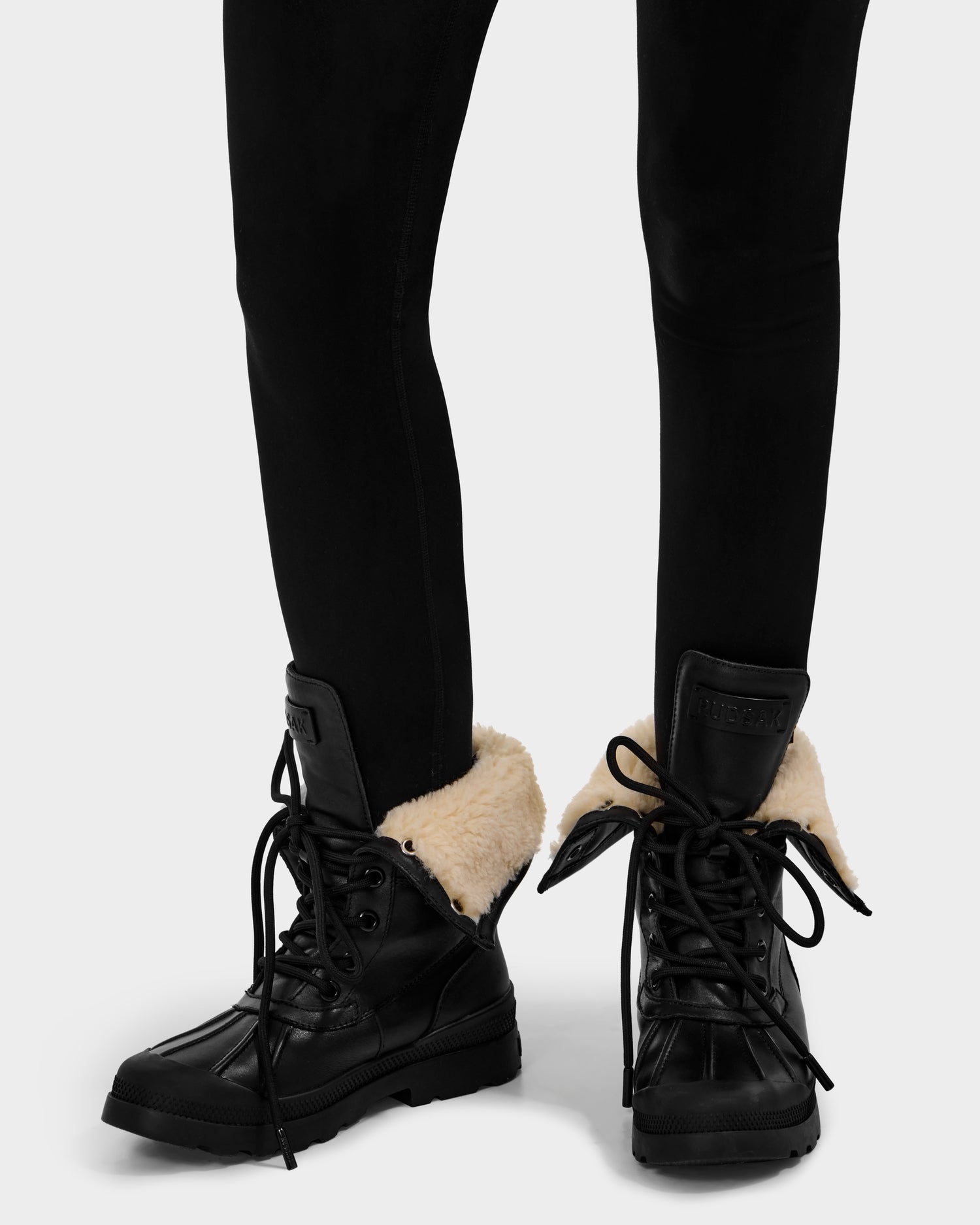 A person wears black pants and Rudsak’s JESI - 8225026 Black winter boots, featuring foldover tops with visible light-colored lining, thick soles, and loosely tied laces for a cozy, stylish look.