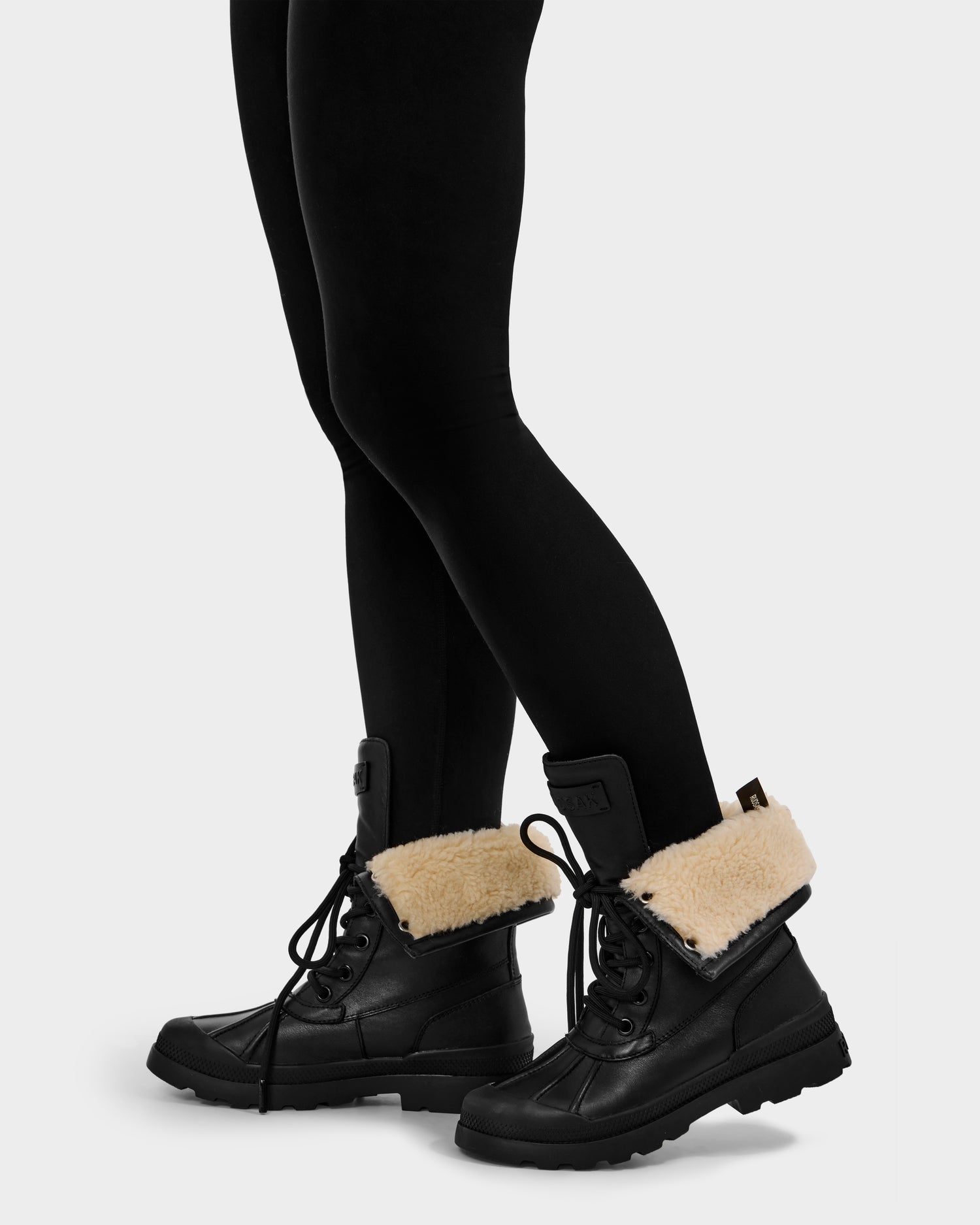 A person wears Rudsak’s JESI - 8225026 Black foldover women’s boots with faux shearling lining and black leggings, standing on a light gray background. The laced-up boots are folded down to showcase the cozy fur.