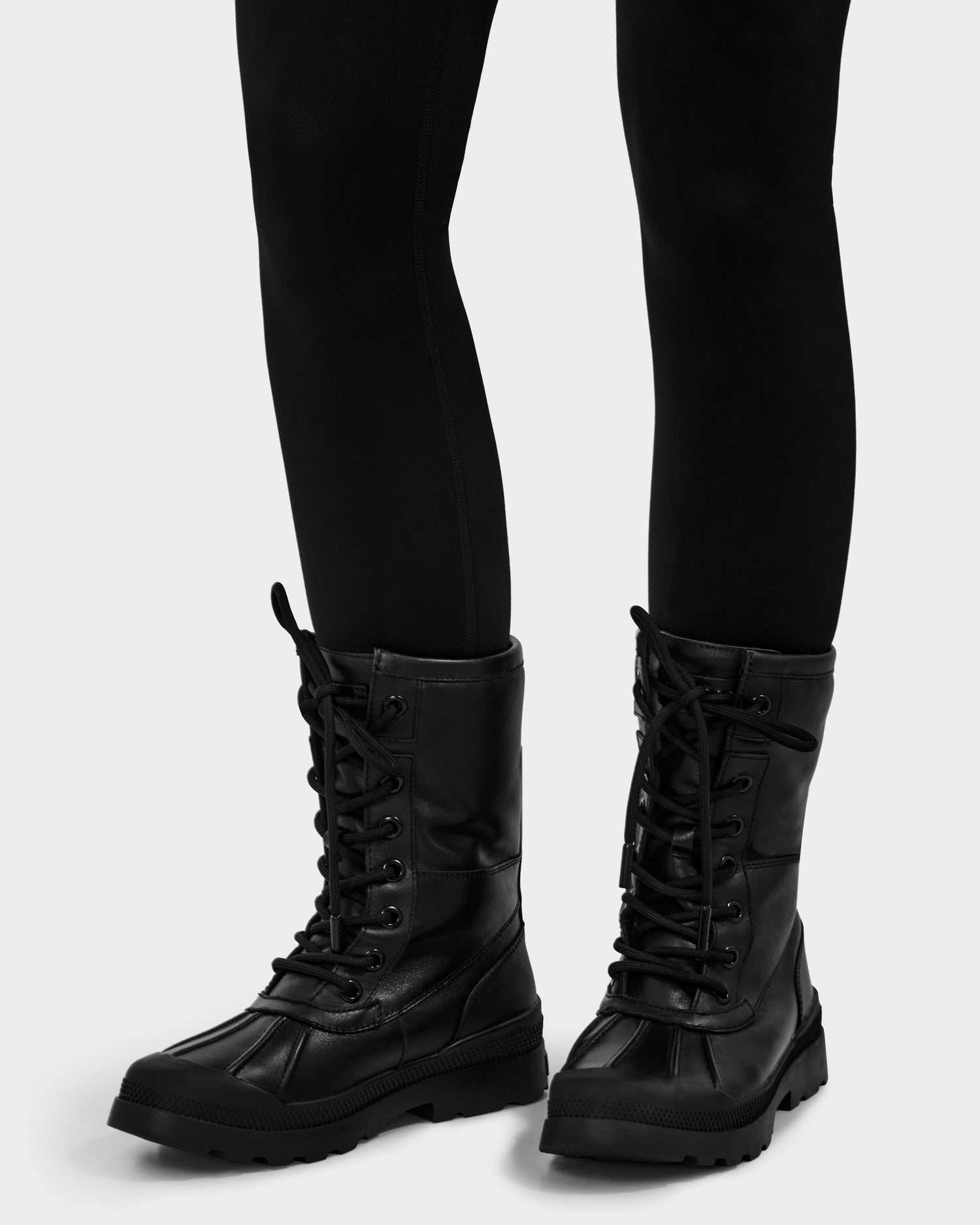 A person wearing Rudsak JESI - 8225026 Black winter-ready combat boots with faux shearling lining and black leggings stands on a light gray surface.