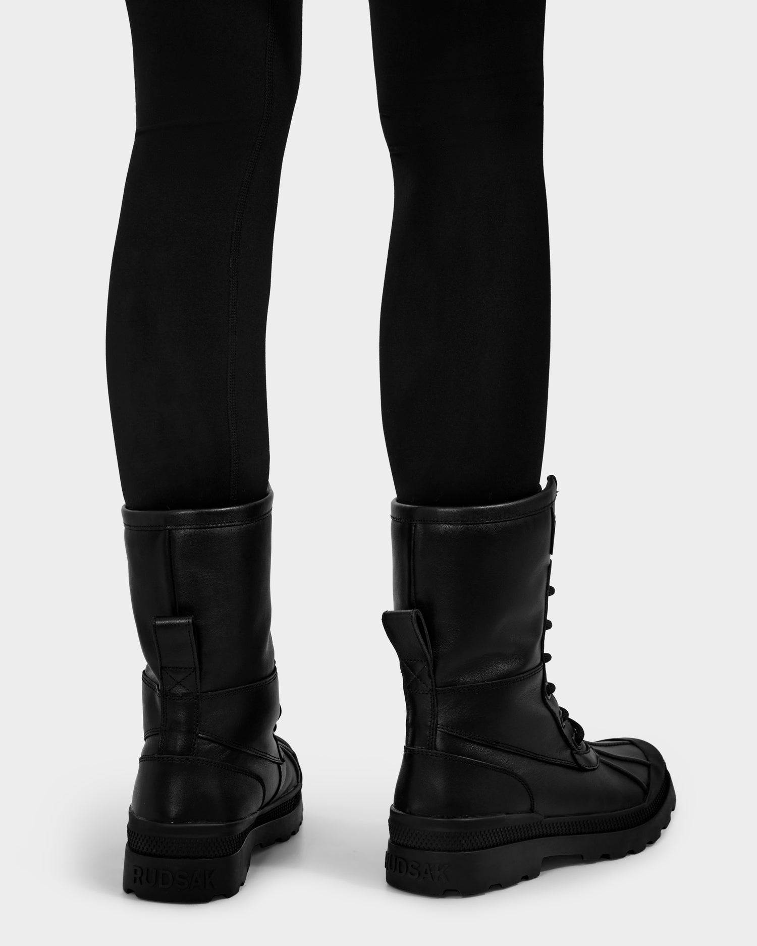 A person shown from behind against a light background wears black leggings and tall black JESI - 8225026 boots by Rudsak, featuring thick winter-ready soles and lace-up details with the brand logo on the heel.