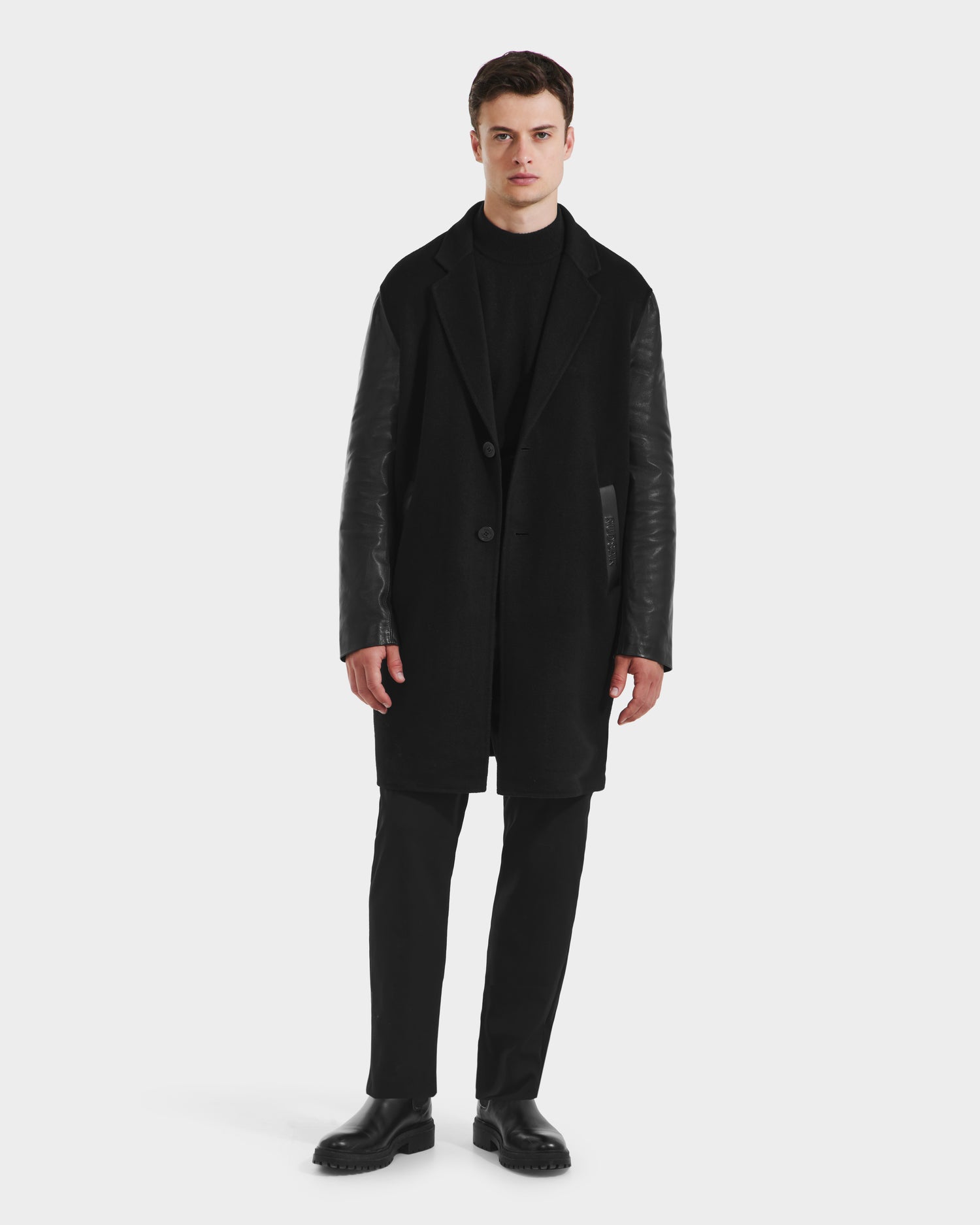 A man stands against a white background wearing the Rudsak JESSE - M125504 Black men’s wool coat with leather sleeves, paired with black pants and shoes. He faces forward neutrally, arms at his sides.