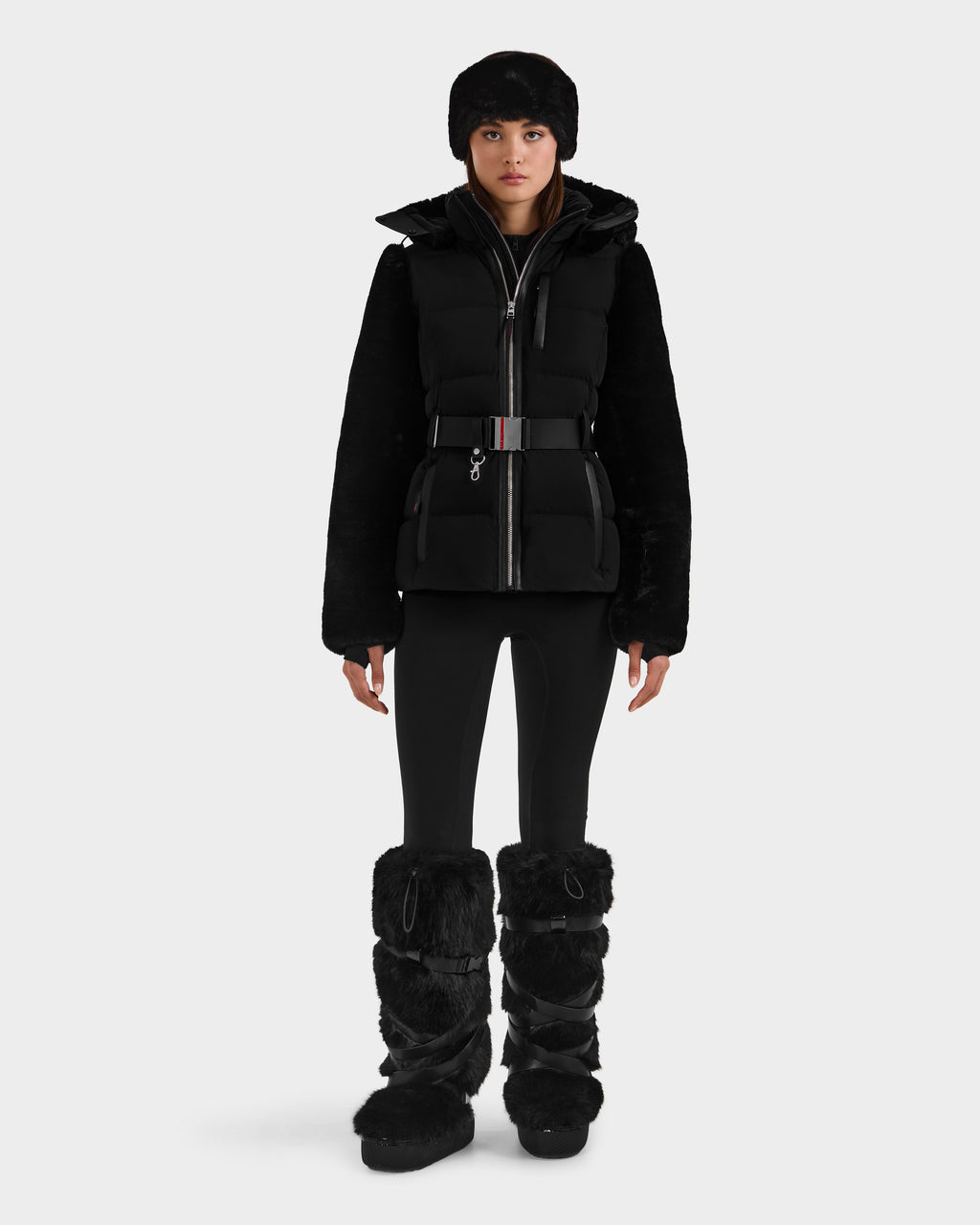A woman models the Rudsak JOANNA F - 8125593 Black winter look—belted puffer jacket, black fur hat, fitted leggings, and tall fur boots—against a white backdrop. Available for pre-order; ships by November 30th.