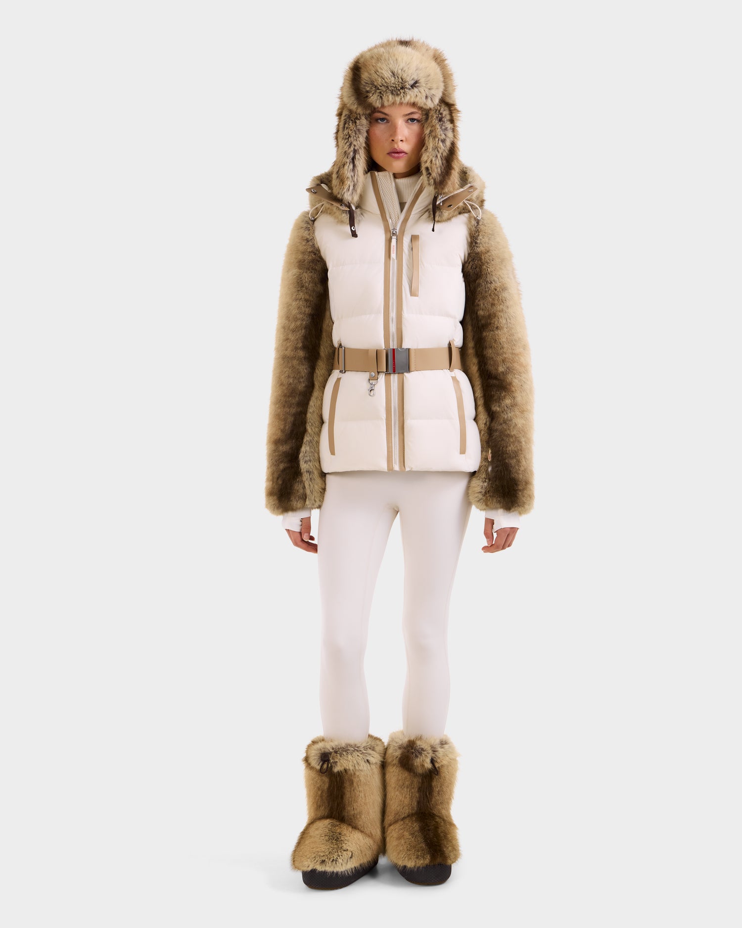 A model in the Rudsak JOANNA F - 8125593 Mocha winter outfit—faux fur sleeves, hat, boots, white leggings, and a belted puffer vest—stands against a white background. Pre-order now; ships November 30th.