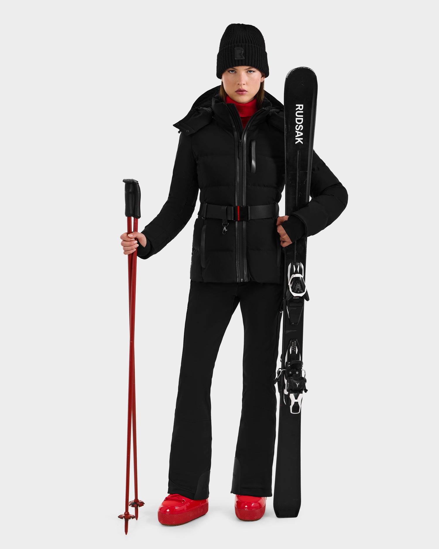A woman in black Rudsak JOANNA - 8124788 ski gear and a beanie holds red ski poles and RUDSAK skis, wearing red boots and a turtleneck. Pre-order now—ships by November 30th.
