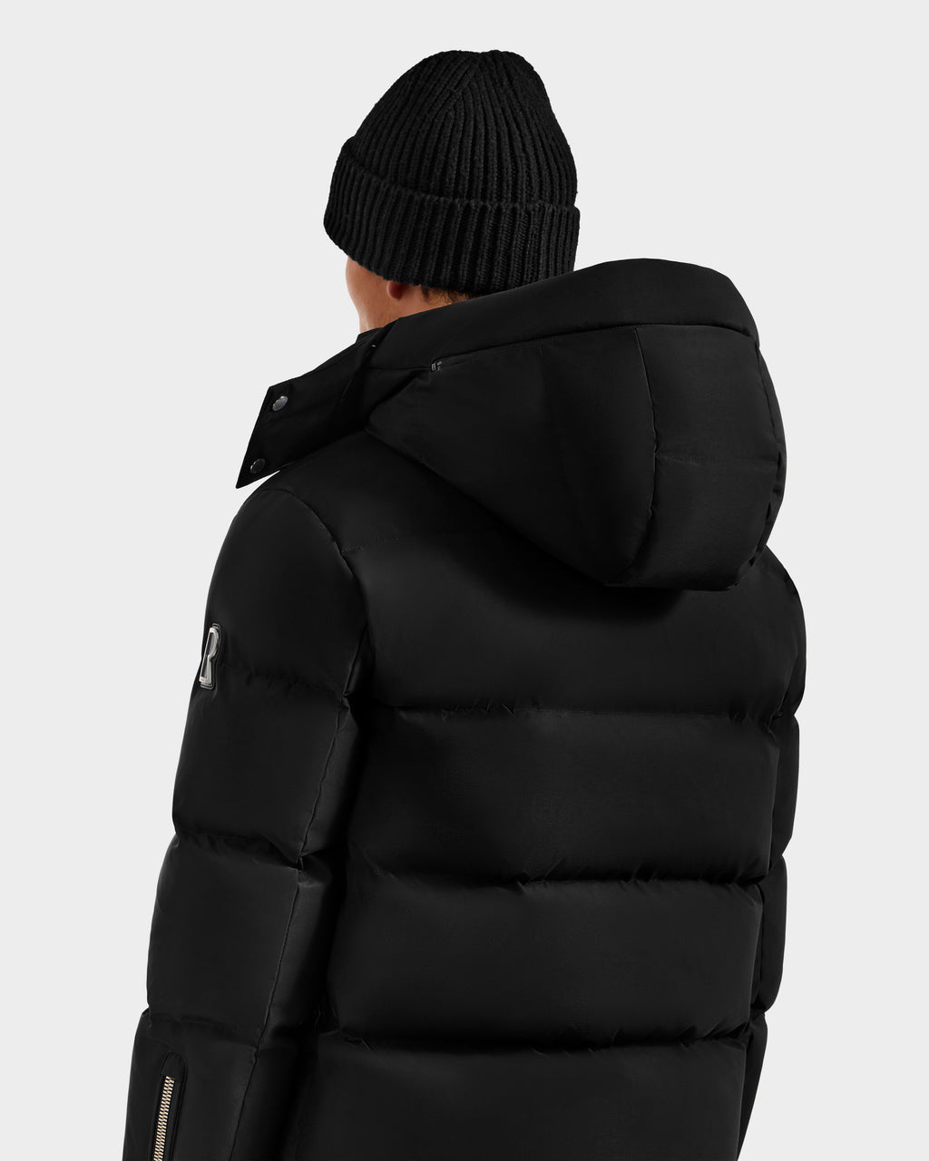 A person wearing the Rudsak JOEL H - 6125582 Black RDS-certified down puffer jacket with a hood and a black knit beanie, shown from the back against a plain light gray background—an understated winter essential.