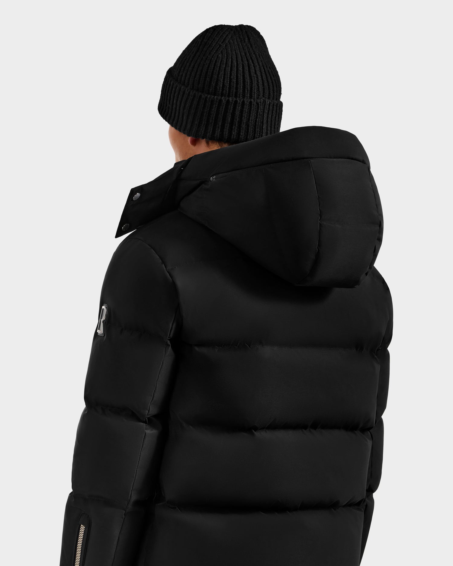 A person wearing the Rudsak JOEL H - 6125582 Black RDS-certified down puffer jacket with a hood and a black knit beanie, shown from the back against a plain light gray background—an understated winter essential.