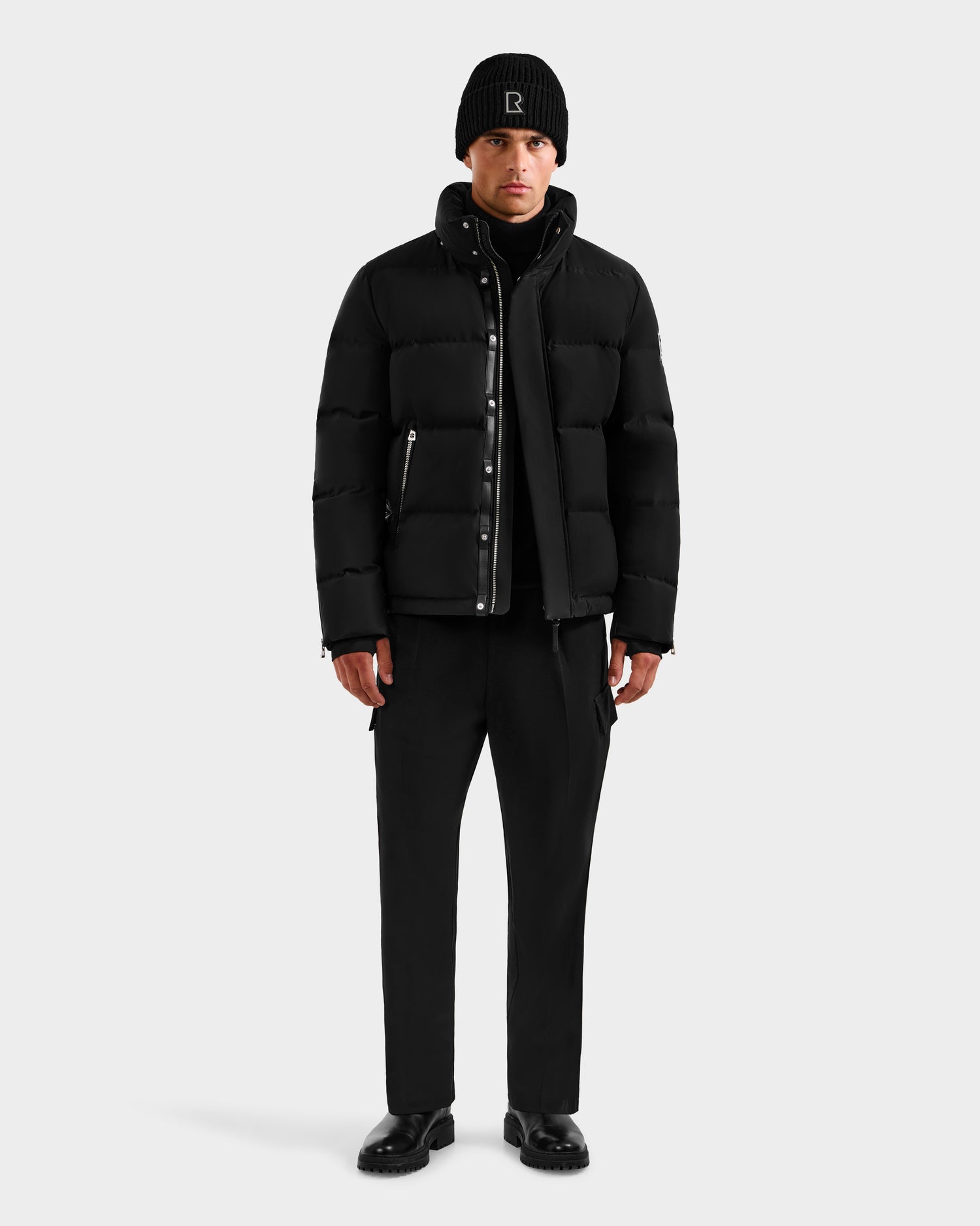 A person stands facing forward in the Rudsak JOEL H - 6125582 Black men’s down parka—an effortlessly cool winter essential—styled with black pants, boots, and a beanie, set against a plain light gray background.