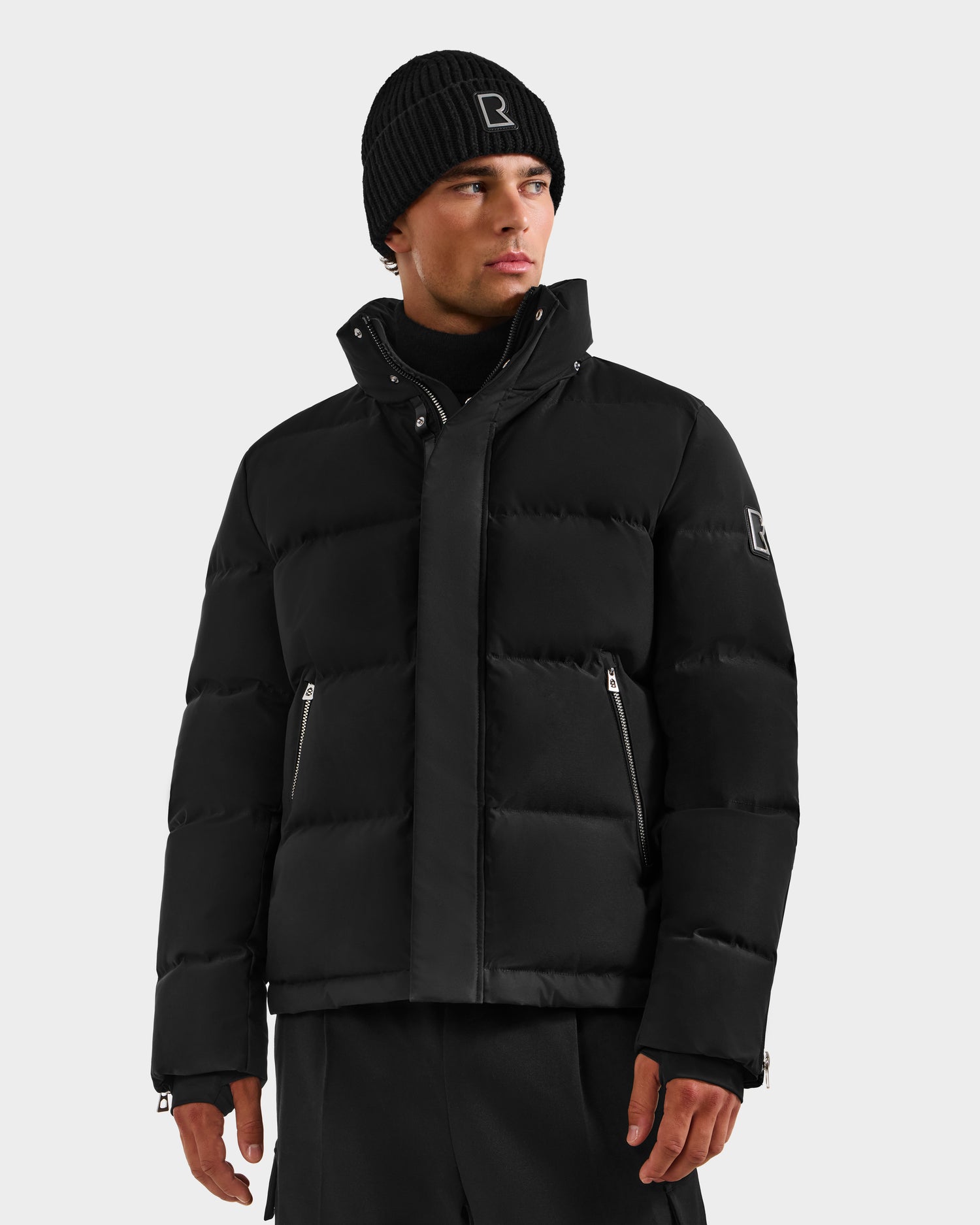 A man wears the Rudsak JOEL H - 6125582 Black men’s down parka with RDS-certified down, a black knit beanie, and black pants, standing against a plain light background—a true winter essential.