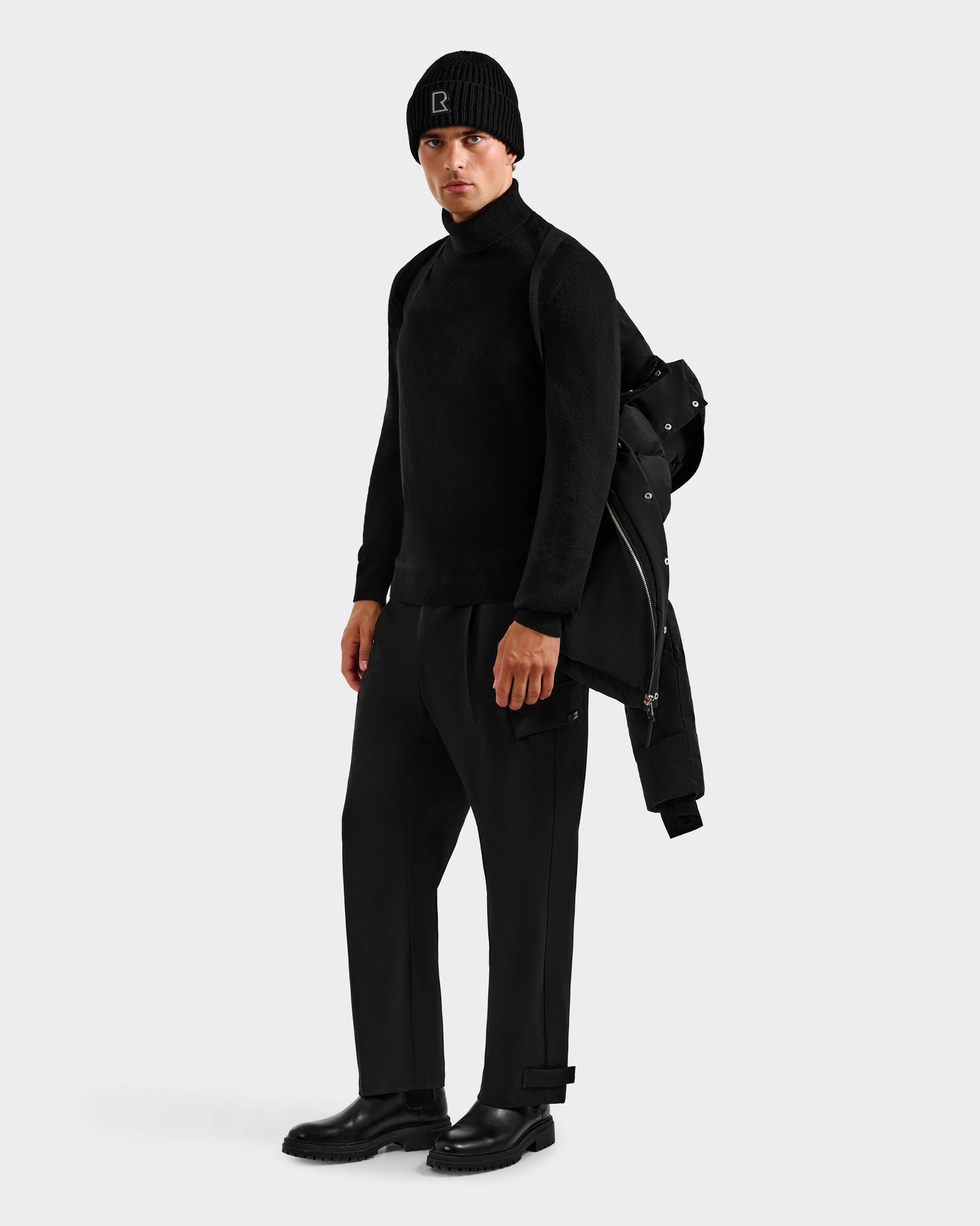 A man stands against a plain background wearing a black beanie, turtleneck, pants, and boots, holding the Rudsak JOEL H - 6125582 Black men’s down parka over his shoulder—a true winter essential.