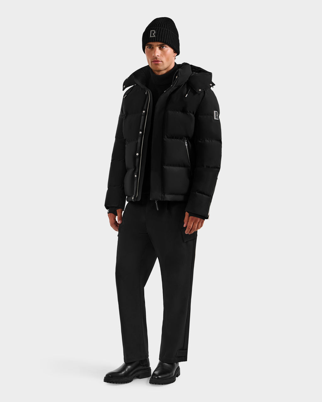 A man stands against a plain background wearing the Rudsak JOEL H - 6125582 Black men’s down parka, paired with a black beanie and pants for a casual, monochromatic winter look crafted with RDS-certified down.