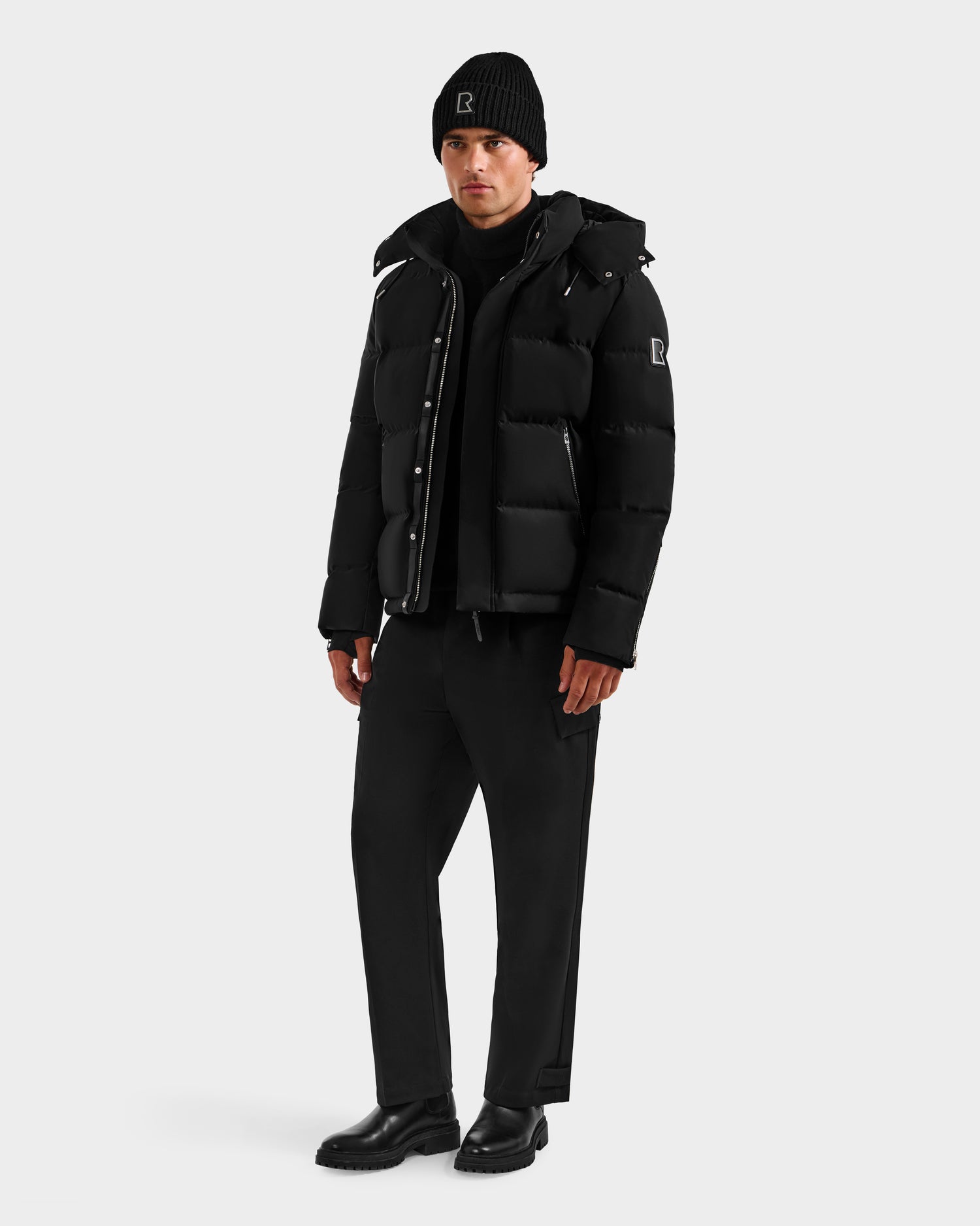A man stands against a plain background wearing the Rudsak JOEL H - 6125582 Black men’s down parka, paired with a black beanie and pants for a casual, monochromatic winter look crafted with RDS-certified down.