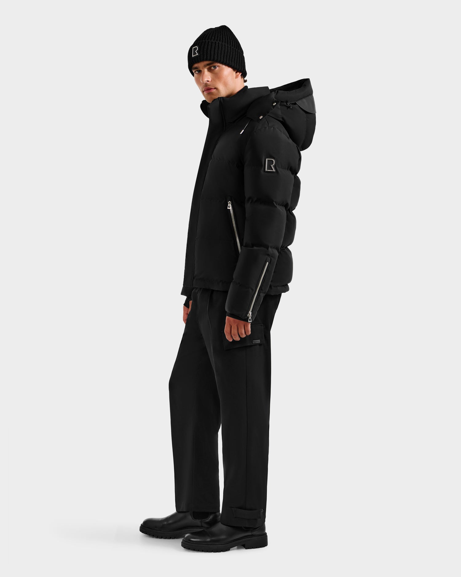 A person in the Rudsak JOEL H - 6125582 Black men’s down parka, paired with black pants, boots, and a beanie, stands against a plain light background—showcasing a sleek winter look while gazing slightly to the side.