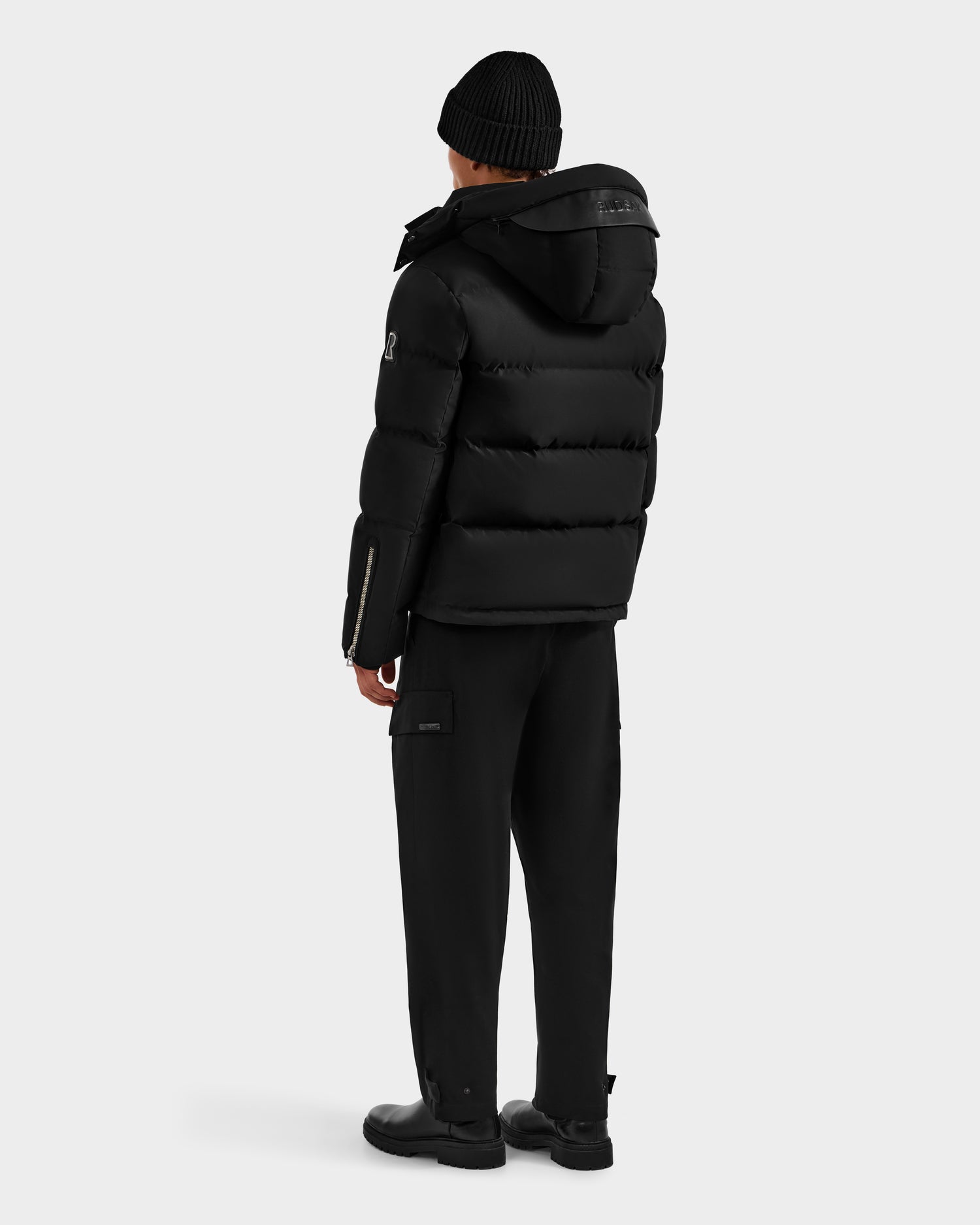 A person in the Rudsak JOEL H - 6125582 Black men’s down parka, black beanie, pants, and shoes stands facing away from the camera against a plain light gray background, showcasing a classic winter essential look.