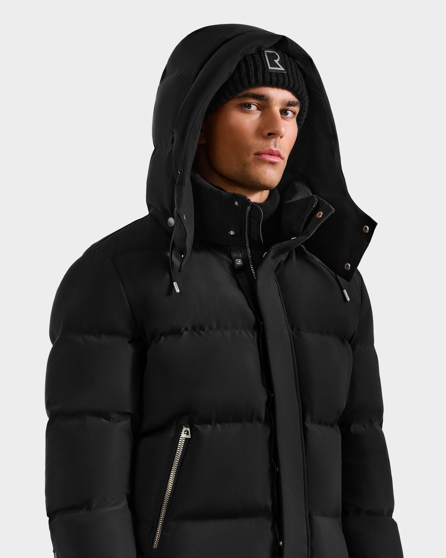 A man models the Rudsak JOEL H - 6125582 Black men’s down parka with hood and a black knit beanie, standing against a plain light background and looking to the side—showcasing this winter essential.