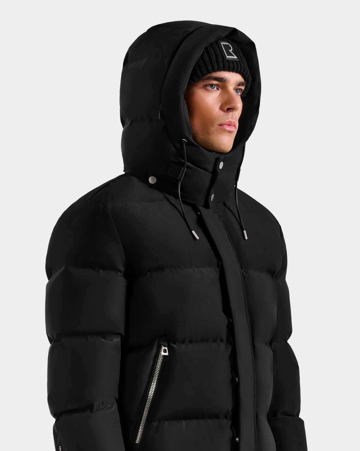 A man wearing the Rudsak JOEL H - 6125582 Black men’s down parka with a hood and a black knit beanie stands against a white background. This winter essential features RDS-certified down, front zipper, and side zippered pocket.