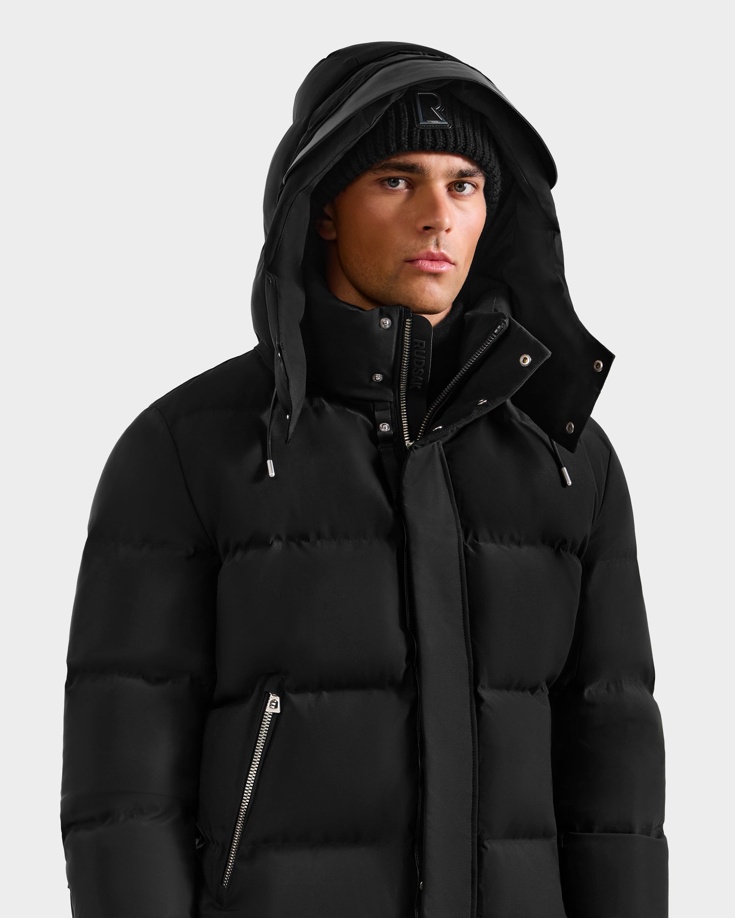 A man wears the JOEL H - 6125582 Black men’s down parka with a hood by Rudsak and a black beanie, looking forward with a neutral expression against a plain light background—the perfect winter essential.