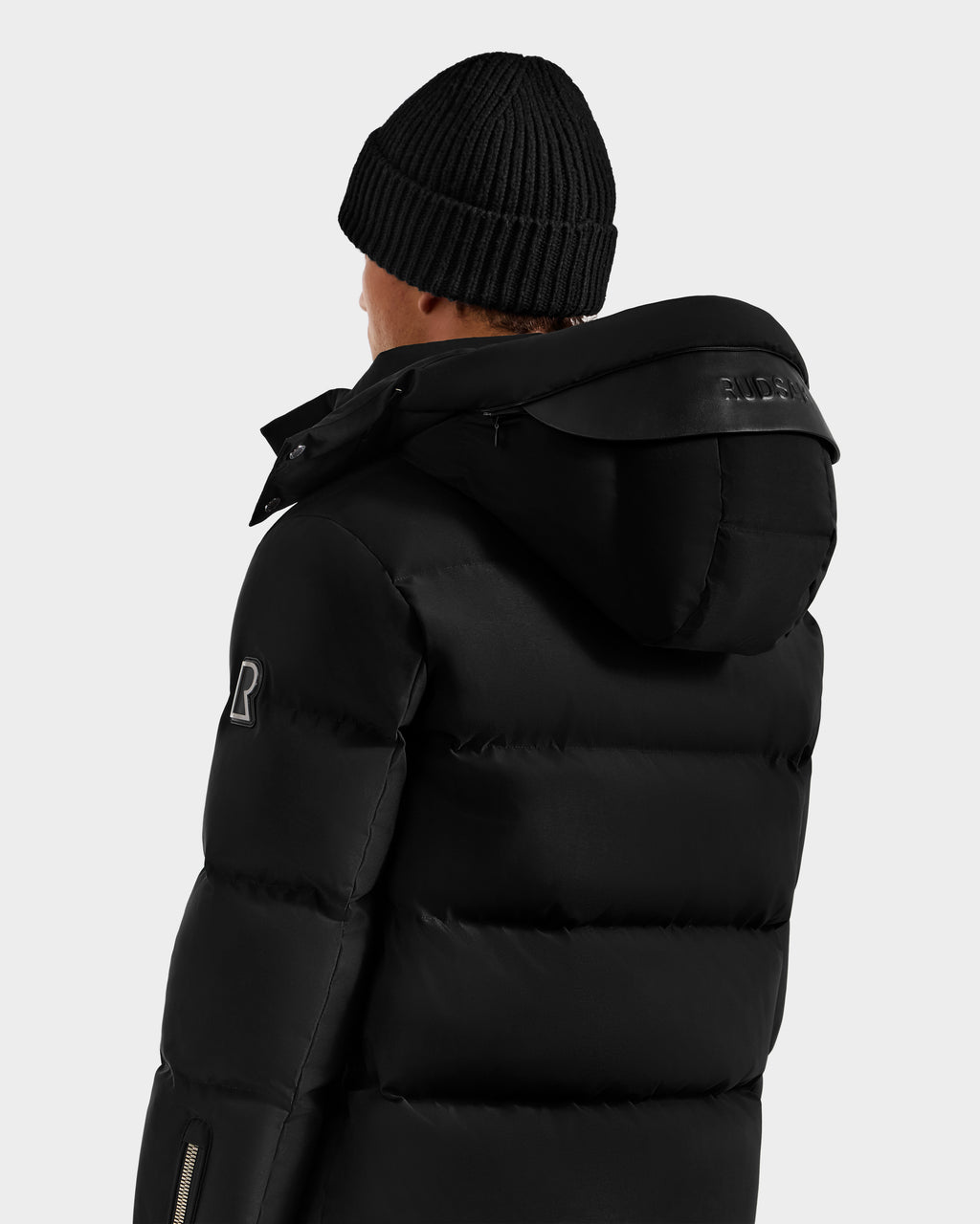 A person wearing the Rudsak JOEL H - 6125582 Black men’s down parka with a hood and a black knit beanie, seen from behind against a plain light background—a true winter essential.
