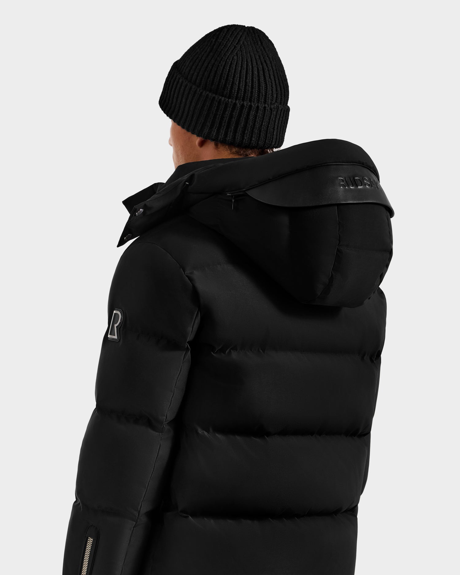 A person wearing the Rudsak JOEL H - 6125582 Black men’s down parka with a hood and a black knit beanie, seen from behind against a plain light background—a true winter essential.