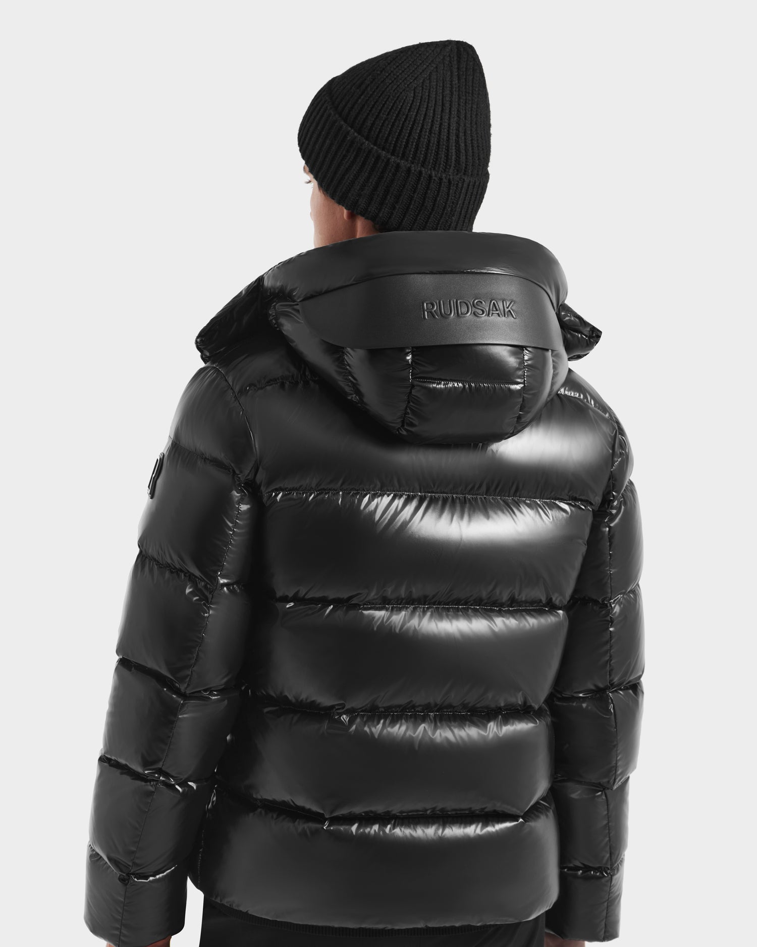 A person wearing the Rudsak JOEL S - 6124606 Black premium down puffer jacket with RUDSAK on the collar and a black knit beanie, facing away from the camera against a plain background—highlighting stylish winter outerwear.
