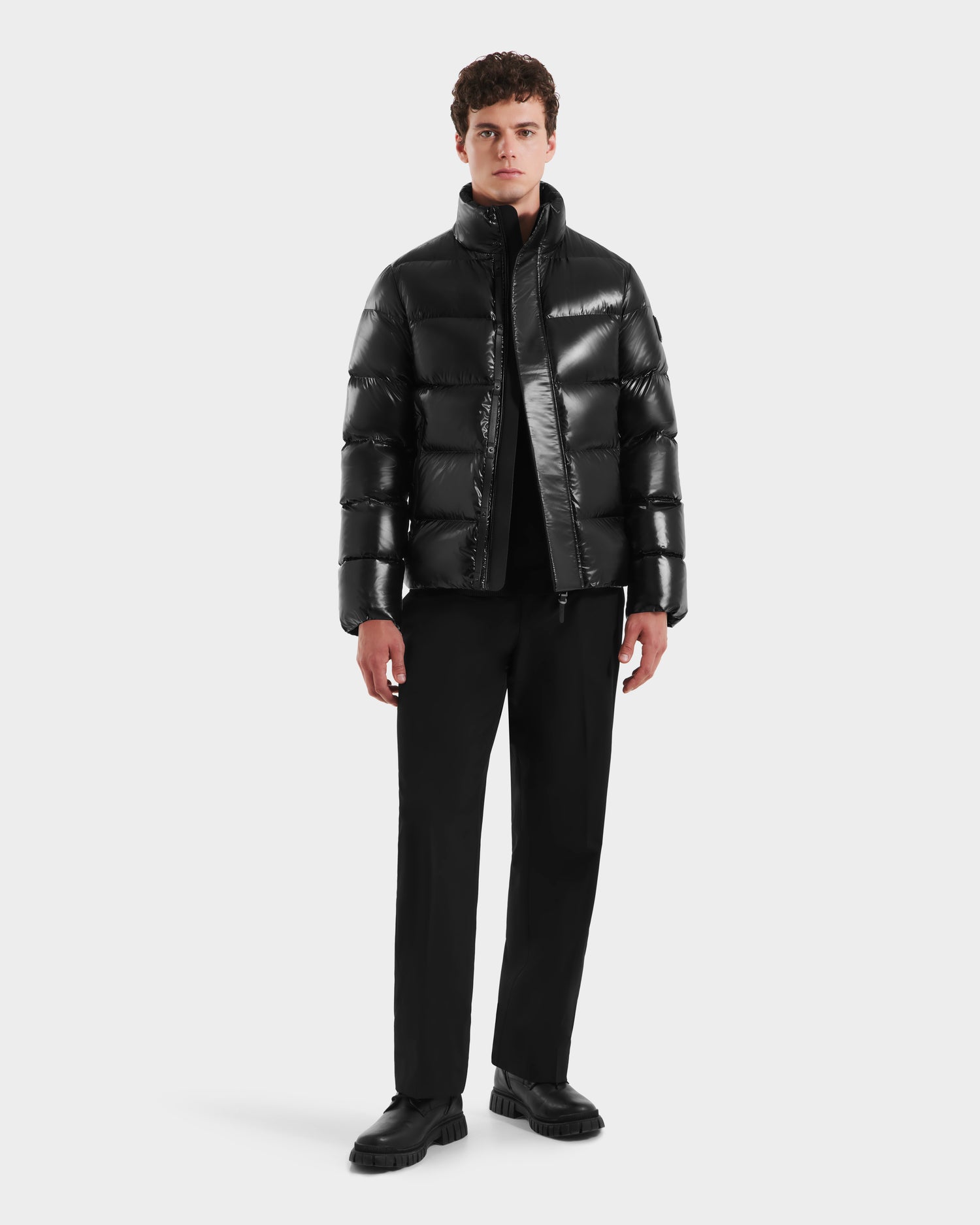 A young man faces forward in a sleek Rudsak JOEL S - 6124606 Black down puffer jacket, paired with black pants, shirt, and shoes, posed against a plain white background.