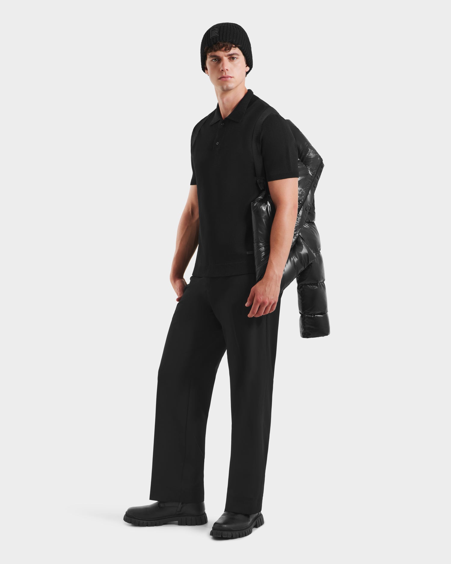 A man stands against a white background, wearing Rudsak’s JOEL S - 6124606 Black premium down puffer jacket draped over his shoulder, paired with a black polo shirt, black pants, shoes, and a knit beanie.