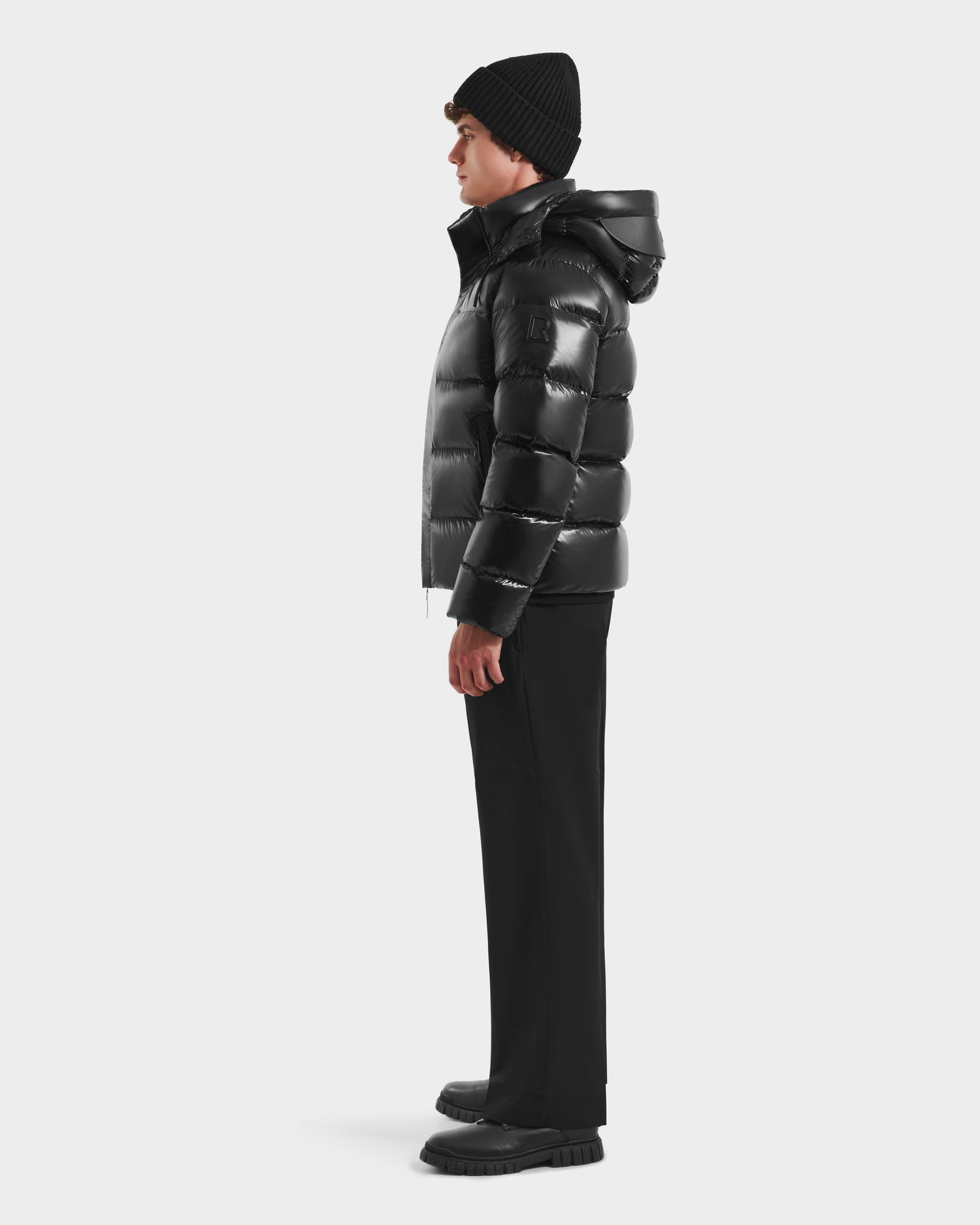 A person in profile wears the Rudsak JOEL S - 6124606 Black premium down puffer jacket, paired with a black beanie, pants, and shoes, standing against a plain white background.