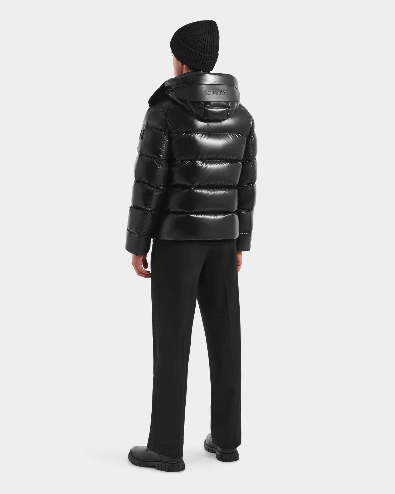A person wearing the Rudsak JOEL S - 6124606 Black down puffer jacket, black pants, shoes, and knit beanie stands facing away against a white background, highlighting premium winter outerwear.