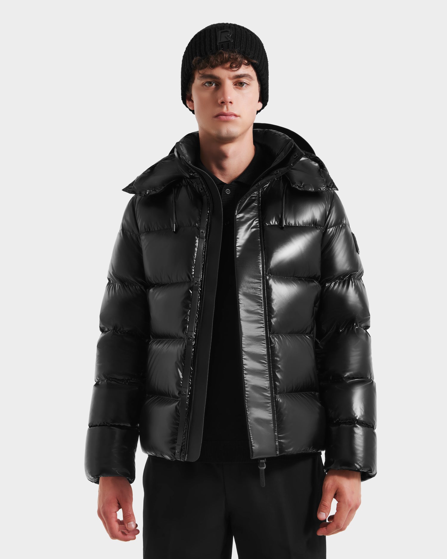 A young man showcases the Rudsak JOEL S - 6124606 Black premium down puffer jacket, paired with a black knit beanie and black pants, standing forward against a plain white background in stylish winter outerwear.