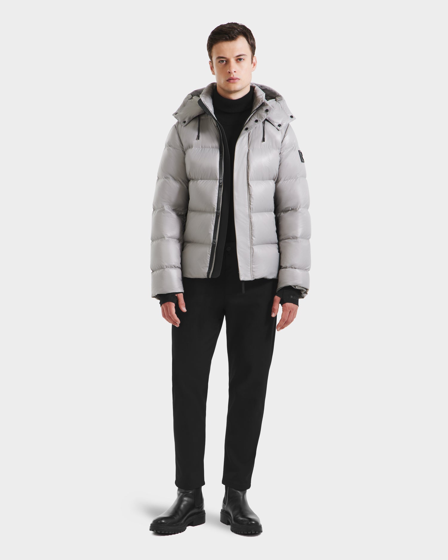 A person faces forward in a Rudsak JOEL S - 6124606 Light Grey premium down puffer jacket, paired with a black turtleneck, pants, and boots—stylish winter outerwear set against a plain white background.